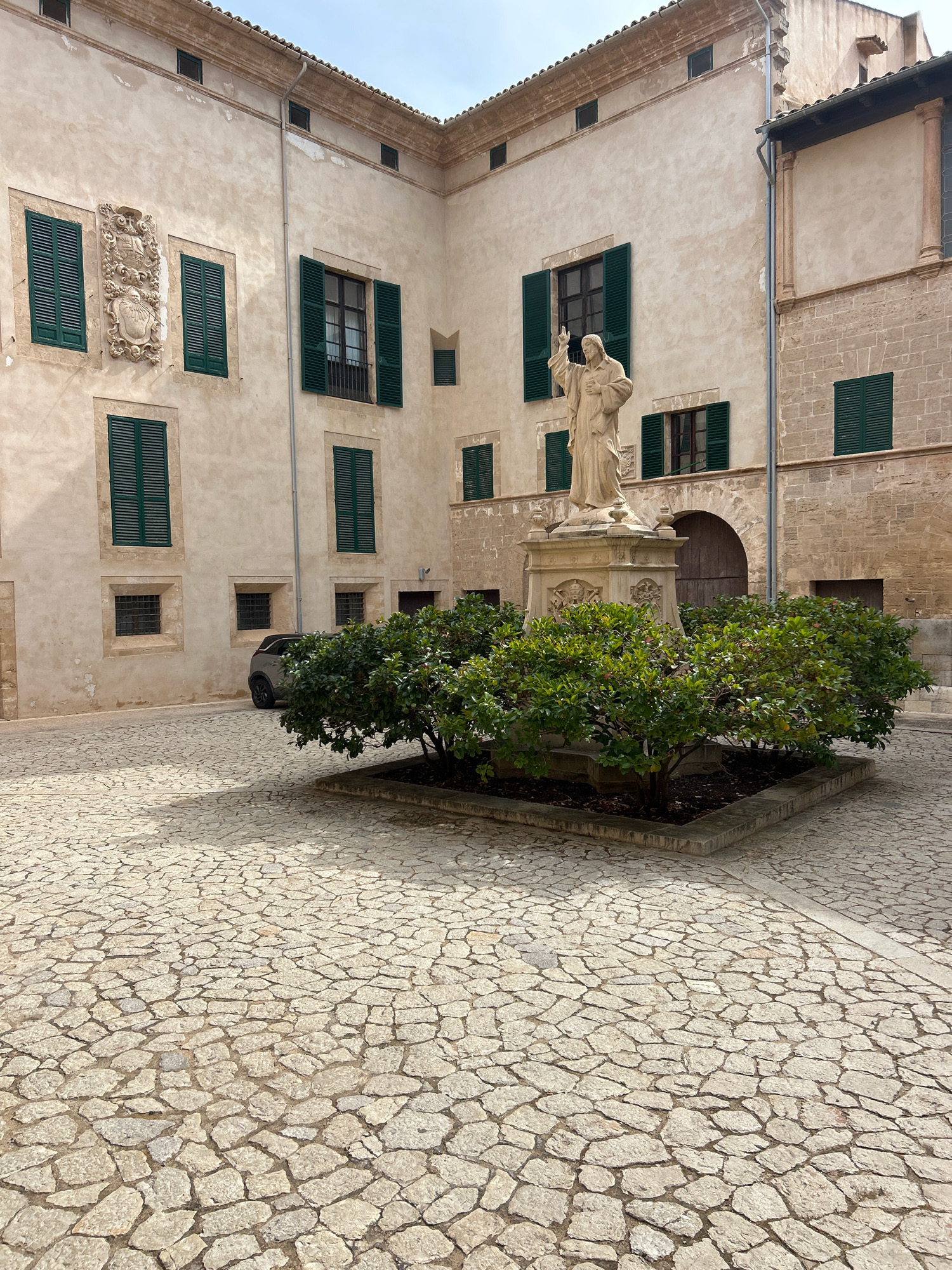 The image depicts a courtyard in Palma de Mallorca, Spain. The courtyard features a cobblestone pavement and is surrounded by buildings with pale stucco walls and green shutters. In the center of the courtyard stands a statue of a man with his right arm raised, wearing a robe. The statue is mounted on a decorative pedestal, and is surrounded by a square planter containing a trimmed green bush. A dark colored car is parked on the left side of the courtyard, partially obscured by the bush. The architecture suggests a historic building, possibly a monastery or a palace. The sky is overcast, indicating a diffused light.