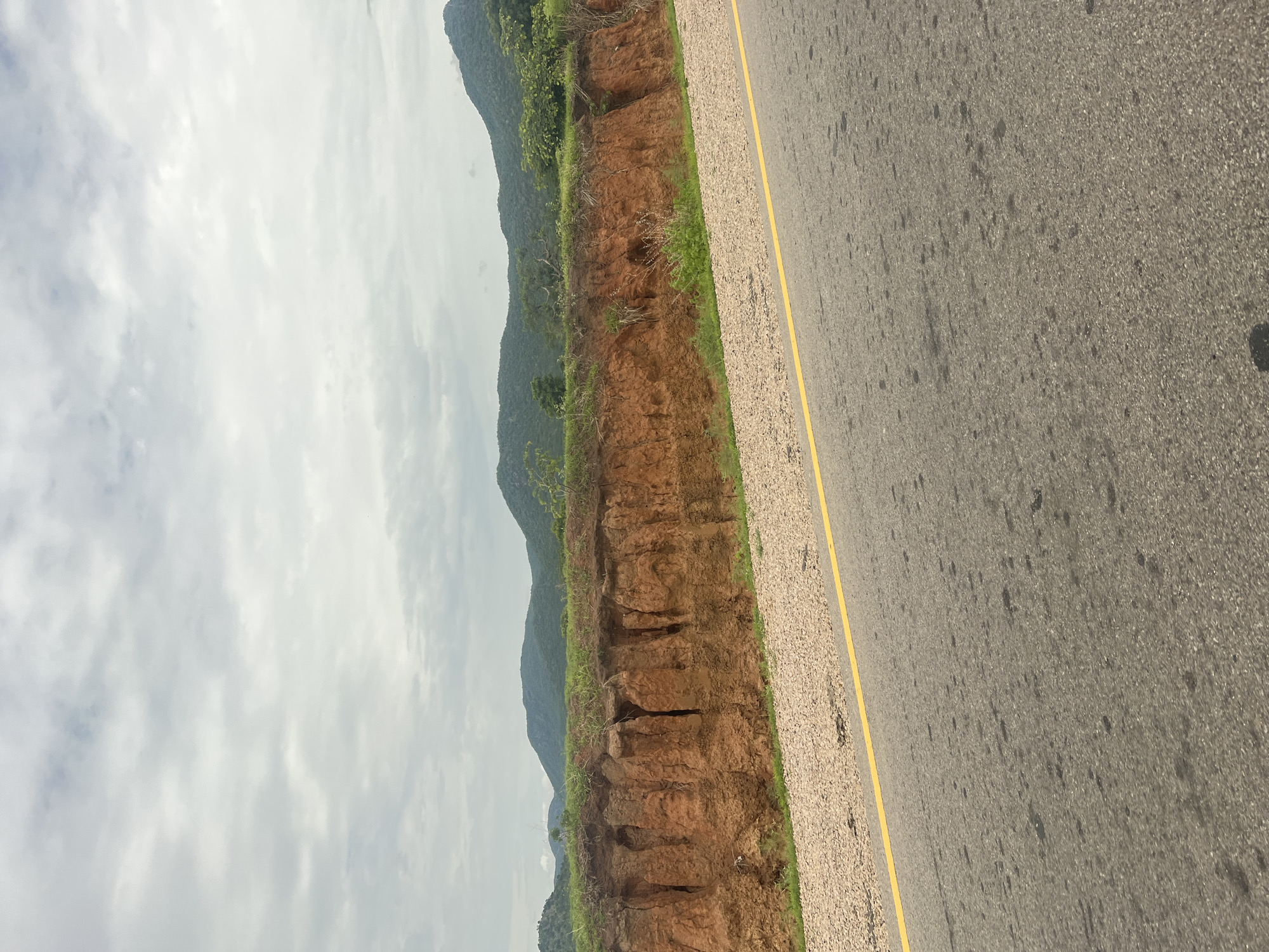 The image shows a road with a yellow line running along the side, bordering a gravel shoulder. Beyond the shoulder, a steep, eroded embankment of red earth is visible, with several deep vertical fissures. The top of the embankment is covered with green grass and some vegetation. In the background, rolling hills covered in lush green trees and vegetation rise under a cloudy sky. The scene appears to be a rural or semi-rural area, possibly along a highway. There are no people or animals visible in the image. The overall impression is one of a natural landscape with signs of erosion.