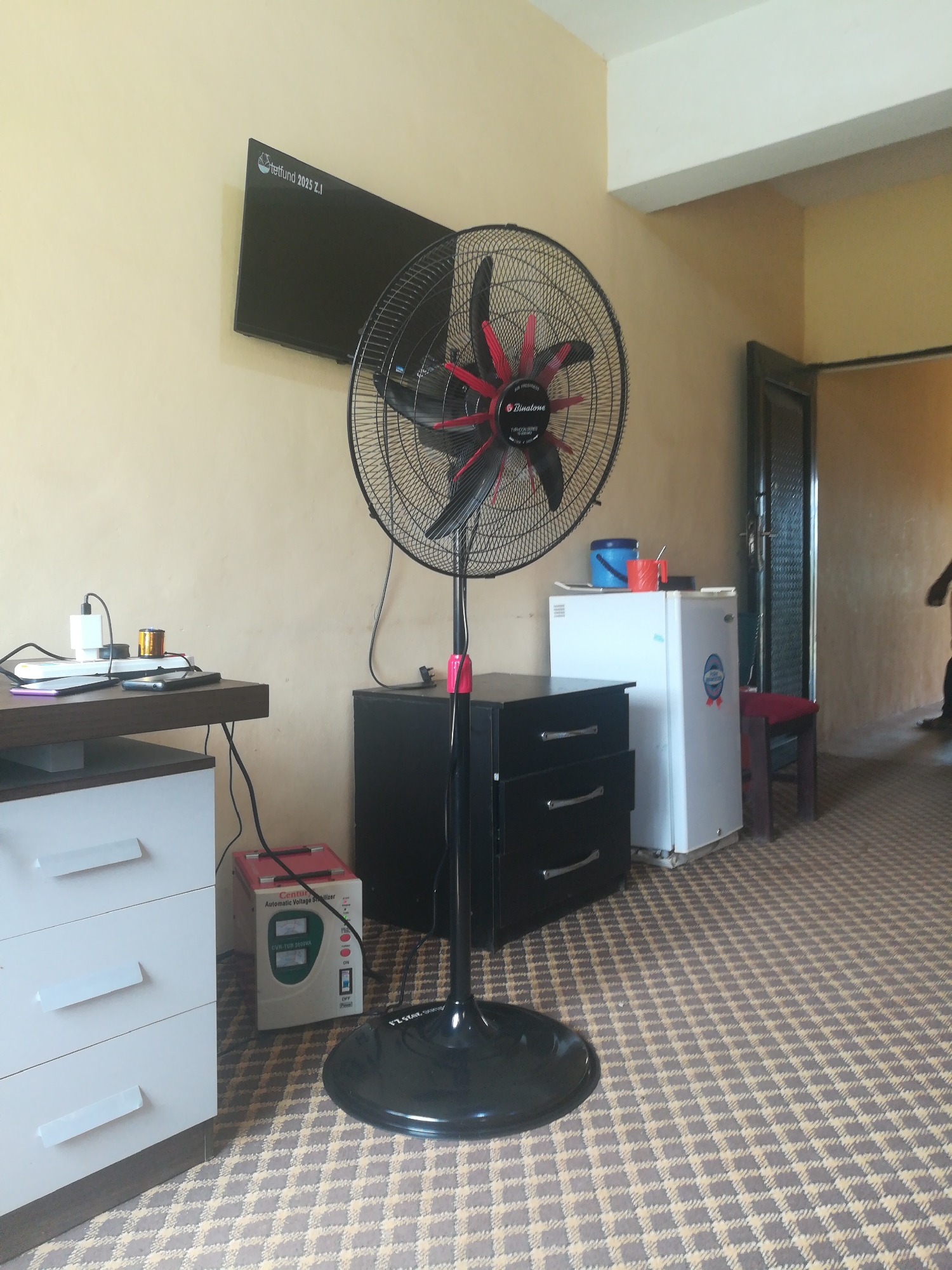 The image depicts an indoor scene in Bisichi, Nigeria, likely a room in a residential or hotel setting. The room is furnished with several objects, including a large black pedestal fan with red accents, a black three-drawer cabinet, a white mini-refrigerator, and a white three-drawer dresser. On top of the dresser, a power strip is visible with a white phone charger and a small cylindrical object, likely a battery or adapter. Two smartphones are placed on the dresser as well.
Adjacent to the dresser and in front of the fan is a white voltage stabilizer with a green digital display. A television is mounted on the wall above the dresser and to the left of the fan. The room has beige walls and a patterned carpet with a geometric design in shades of brown and beige.
To the right of the refrigerator, a doorway with a dark-framed door is visible, leading to another area. A small wooden stool with a red cushion sits near the doorway. In the background, a person is partially visible, walking away from the camera, suggesting activity within the premises. The lighting suggests it is daytime, with no indication of specific weather conditions.
Visible text includes "tettfund 2025 Z.I" on the television screen and "Binatone" on the fan. The voltage stabilizer also has text on its display, though it is not fully legible. The overall scene conveys a functional, somewhat cluttered living or working space.