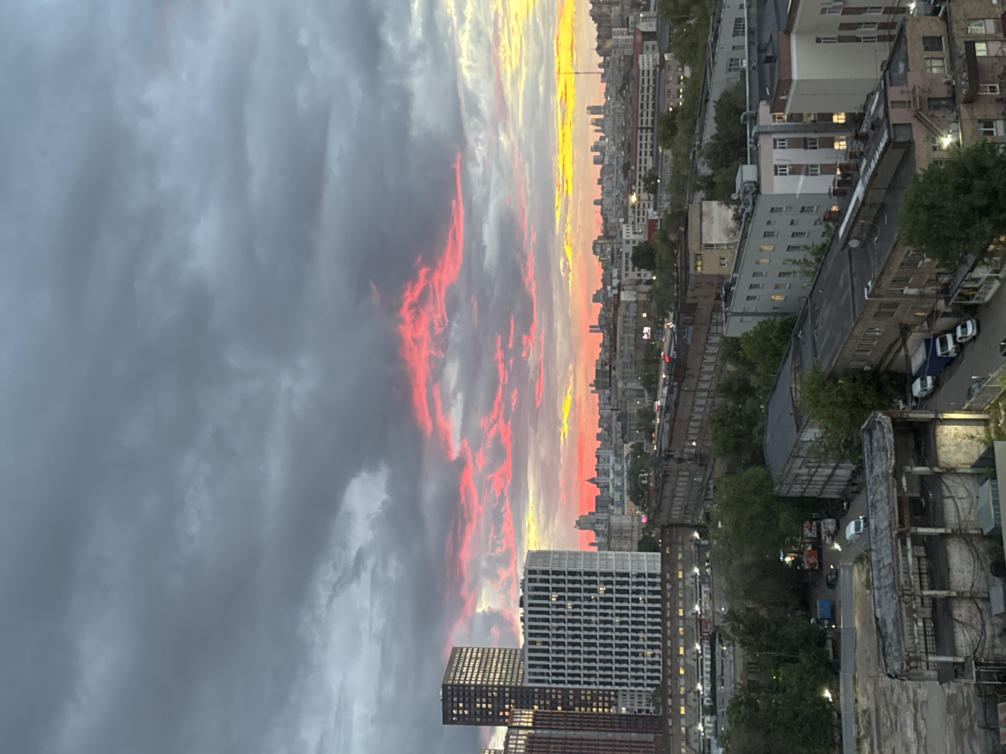 The image depicts a cityscape at dusk or dawn, likely in Moscow, Russia. The sky is dramatic with dark grey clouds dominating the left side, transitioning to a vibrant mix of red, orange, and yellow near the horizon on the right. This colorful band indicates the presence of the sun just below the skyline.
Below the sky, a densely populated urban area stretches towards the horizon. Numerous buildings of varying heights and architectural styles are visible. Some are taller modern structures with grid-like facades, while others are older, more traditional buildings. Lights are on in many of the buildings, suggesting it is either early morning or evening.
The streets below are lined with trees and appear to have vehicle traffic, indicated by the faint streaks of light from headlights. Infrastructure, like bridges or elevated walkways, is also present. The image is taken from a high vantage point, possibly from a tall building or apartment complex, offering a panoramic view of the city and the sky.