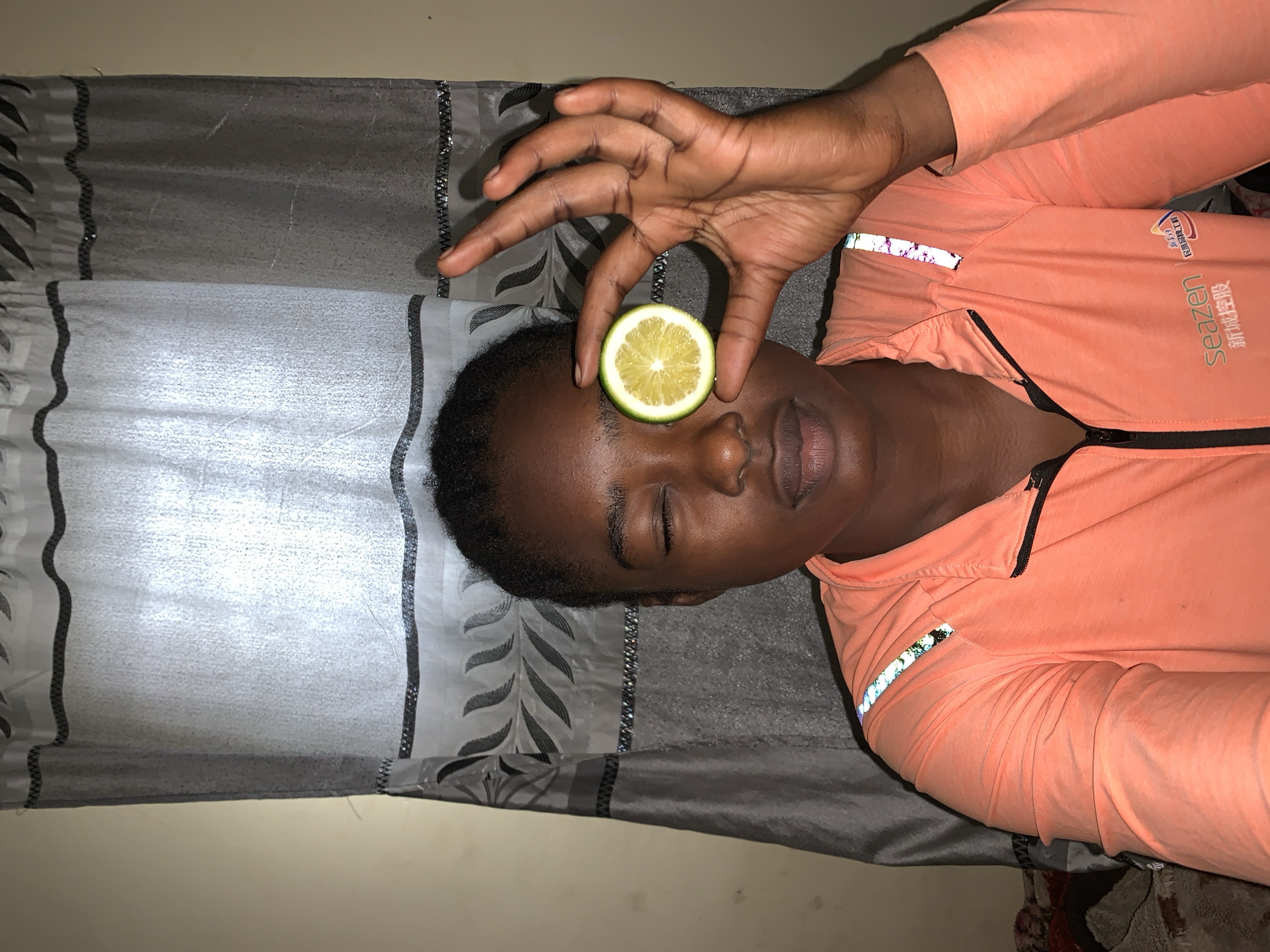 A person is lying on their side against a gray curtain with a leaf pattern. The person is wearing a salmon-colored long-sleeved shirt with a black zipper. They have dark skin and closed eyes. A slice of lemon is positioned between their fingers above their closed eyes.
There is no evidence in the image to support this being in Rafin Par, Nigeria.