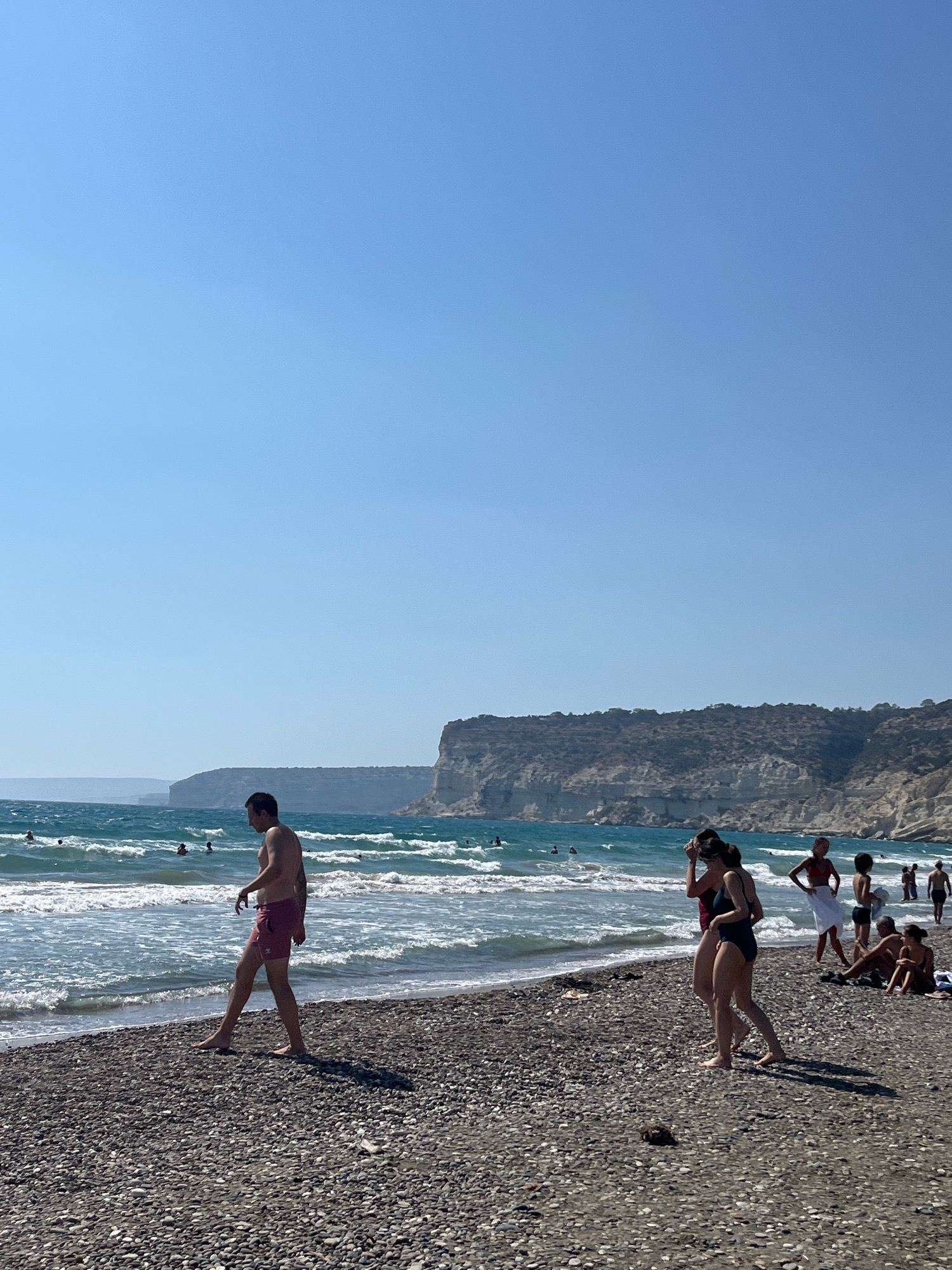 A bright, sunny day at a pebbled beach with people enjoying the water and shoreline. The ocean is a vibrant turquoise with moderate waves. In the foreground, a man in red swim trunks walks along the beach. Further back, several other individuals are visible, some in the water and others relaxing on the shore. Behind the beach, rugged cliffs rise steeply, with a clear blue sky overhead. The location is identified as Episkopi beach, Episkopi, Limassol District, Cyprus.