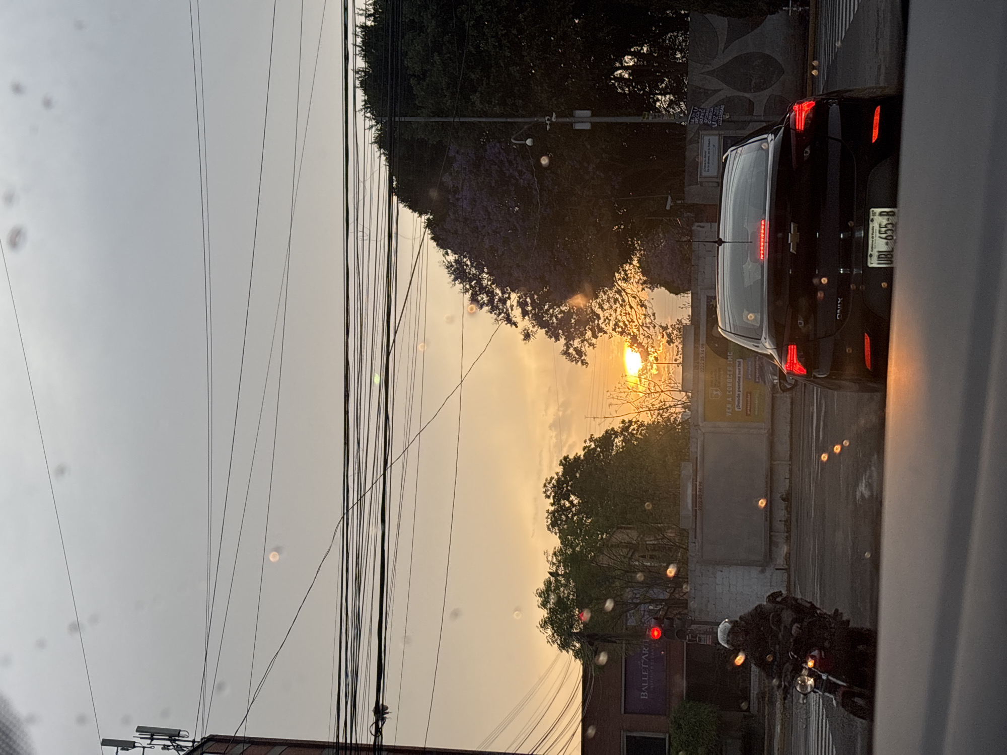The image captures a scene in Puebla, Mexico, at what appears to be sunset or sunrise. The sky is a gradient of grey to orange. A web of overhead power lines is visible. A black car is on the right, partially blocking the view, with a license plate number UBL-655-B visible. The tail lights of the car are illuminated. Next to the car, on the right side of the street, there is a motorcyclist. The street appears wet. Across the street, there are buildings, trees and a mural. A sign with "BALLET ART INSTITUTO DE DAN" can be seen.