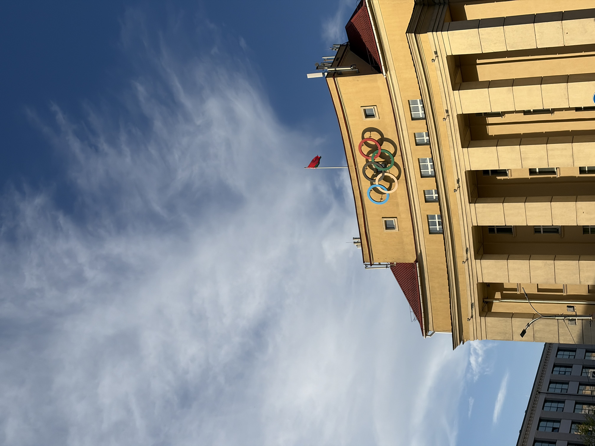The image shows a building in Minsk, Belarus. The building is yellow and has the Olympic rings on the side. There is a red flag on a flagpole. The sky is blue with white clouds. Another building with an interesting design is in the background.