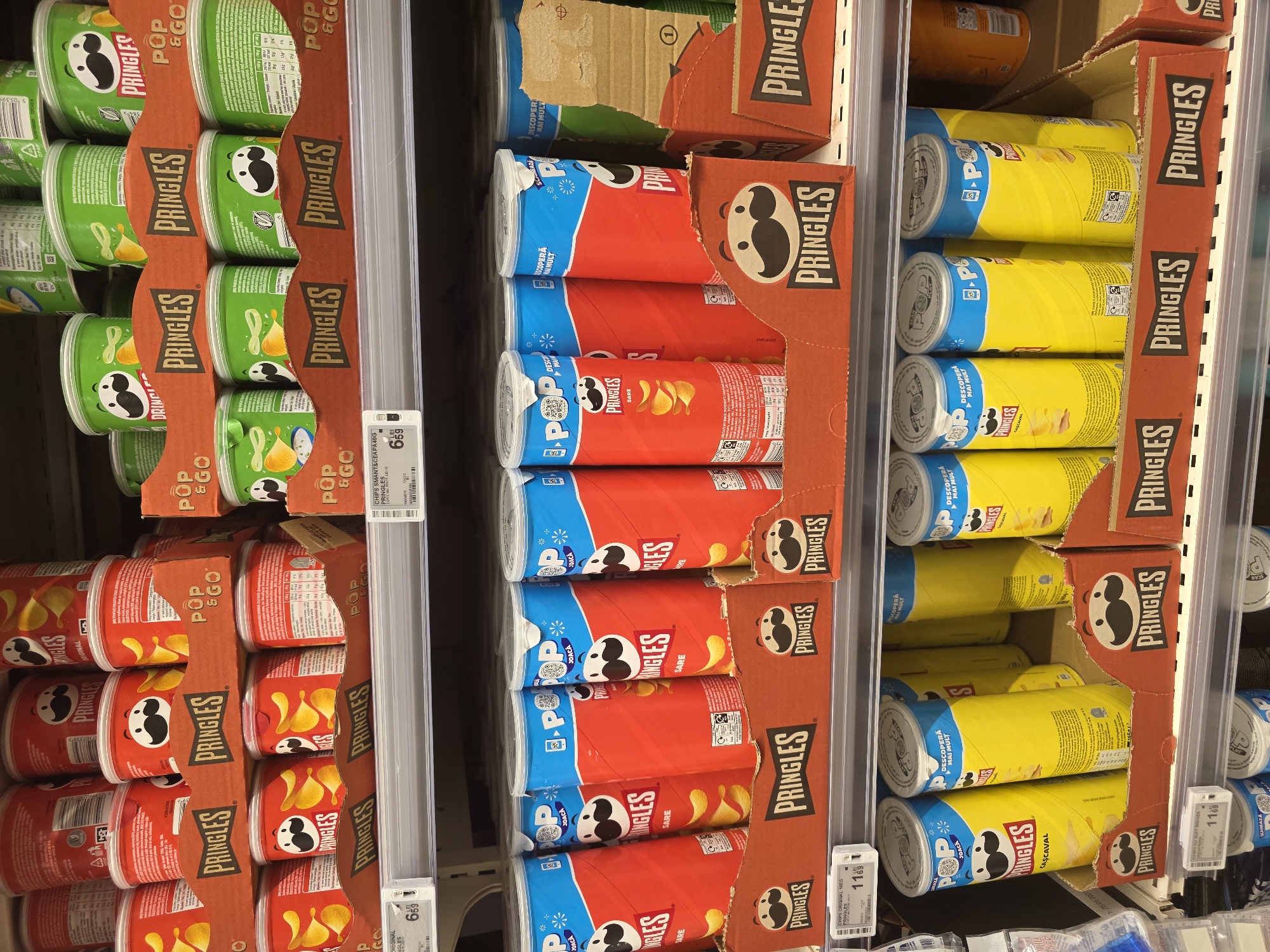 The image displays a retail store shelf stocked with multiple stacks of Pringles potato chip cans. The Pringles cans vary in flavor and color, with prominent red cans featuring the original flavor and yellow cans likely representing cheese flavor. They are arranged in cardboard display trays. Some of the cans have "POP & GO" branding on them. Price tags are visible on the shelves, indicating prices in "LEI". The environment appears to be a supermarket aisle, possibly in Bucharest, Romania, given the currency. There are no people visible in the frame. The lighting suggests an indoor setting. The overall scene is one of product display and retail merchandise.