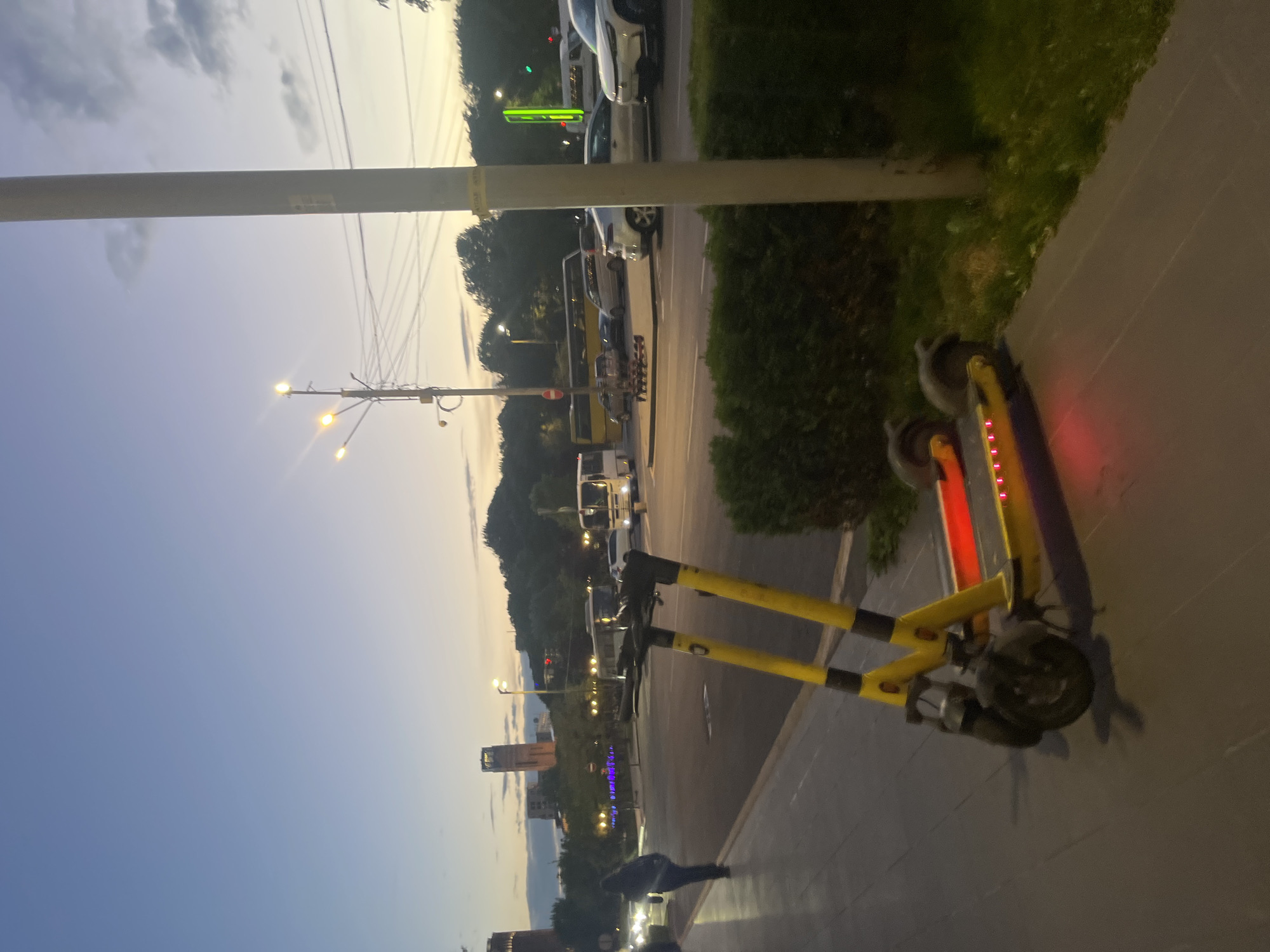 The image shows a partially obscured view of Yerevan, Armenia, at dusk. A yellow and black electric scooter is parked on a sidewalk. A street with car traffic runs adjacent to the sidewalk. A person is walking in the background. In the distance, a building is visible. Streetlights are on.