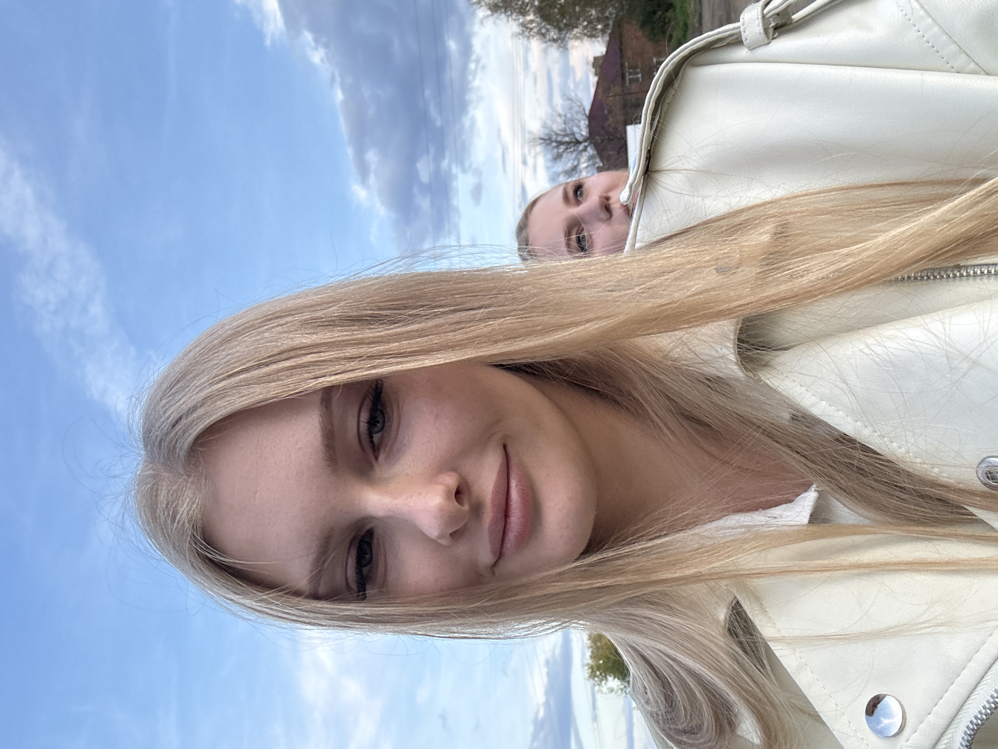 An outdoor, eye-level selfie shows two young Caucasian women in the foreground and middle ground, with a background of a cloudy sky, trees, and a building. The woman in the foreground, who is closer to the camera and slightly to the left, has long blonde hair that falls over her left shoulder and down her chest. She is wearing a white shirt and a light-colored leather jacket. Her head is tilted to the right, and she is looking at the camera with a slight smile. She has blonde eyebrows and dark eyelashes. The woman in the middle ground, who is slightly to the right of the foreground woman and farther from the camera, also has blonde hair. She is looking to the right, away from the camera. She is wearing a white shirt. The background consists of a pale blue sky with scattered clouds. To the right of the middle ground woman, there is a building with a red roof and some trees. The overall lighting suggests it is daytime, possibly late afternoon or early evening.