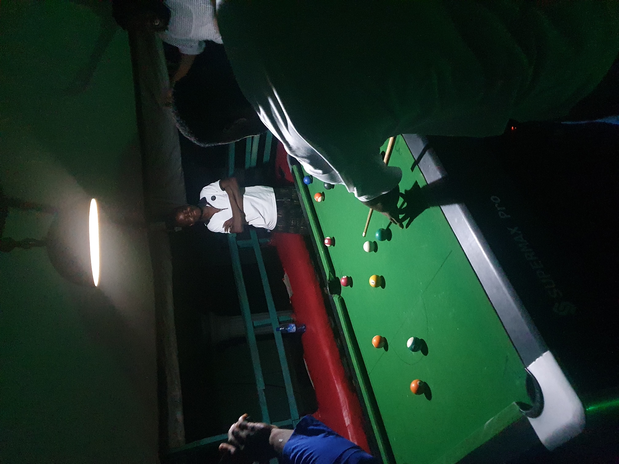 A group of young men are playing pool in a dimly lit indoor setting. The pool table is green with various colored balls scattered across it, some in pockets and some on the table. One person is actively playing, leaning over the table with a cue stick. Another person stands with their arms crossed, observing the game. A third person, wearing a white shirt, is partially visible in the background. The environment appears to be a casual establishment, possibly a bar or a recreational center, with a ceiling lamp providing overhead illumination. The overall atmosphere suggests a friendly game amongst friends. The scene is located in Lokoja, Nigeria.