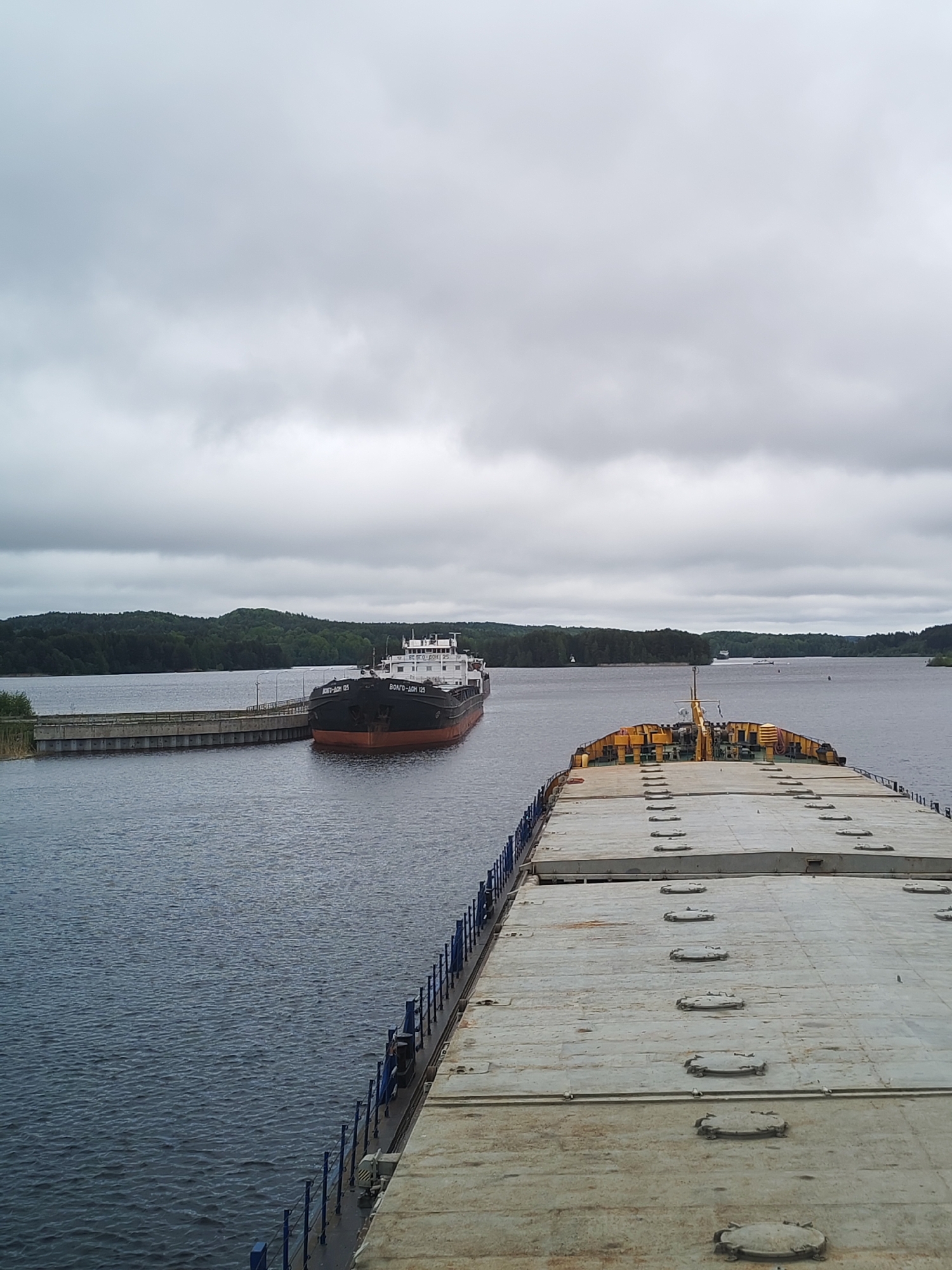 The image depicts a waterway near Белоусово, Russia under an overcast sky. A large, flat-deck barge occupies the foreground with a second, smaller cargo ship docked alongside a concrete pier in the middle distance. Trees line the shoreline in the background. The water is calm with a slight ripple.