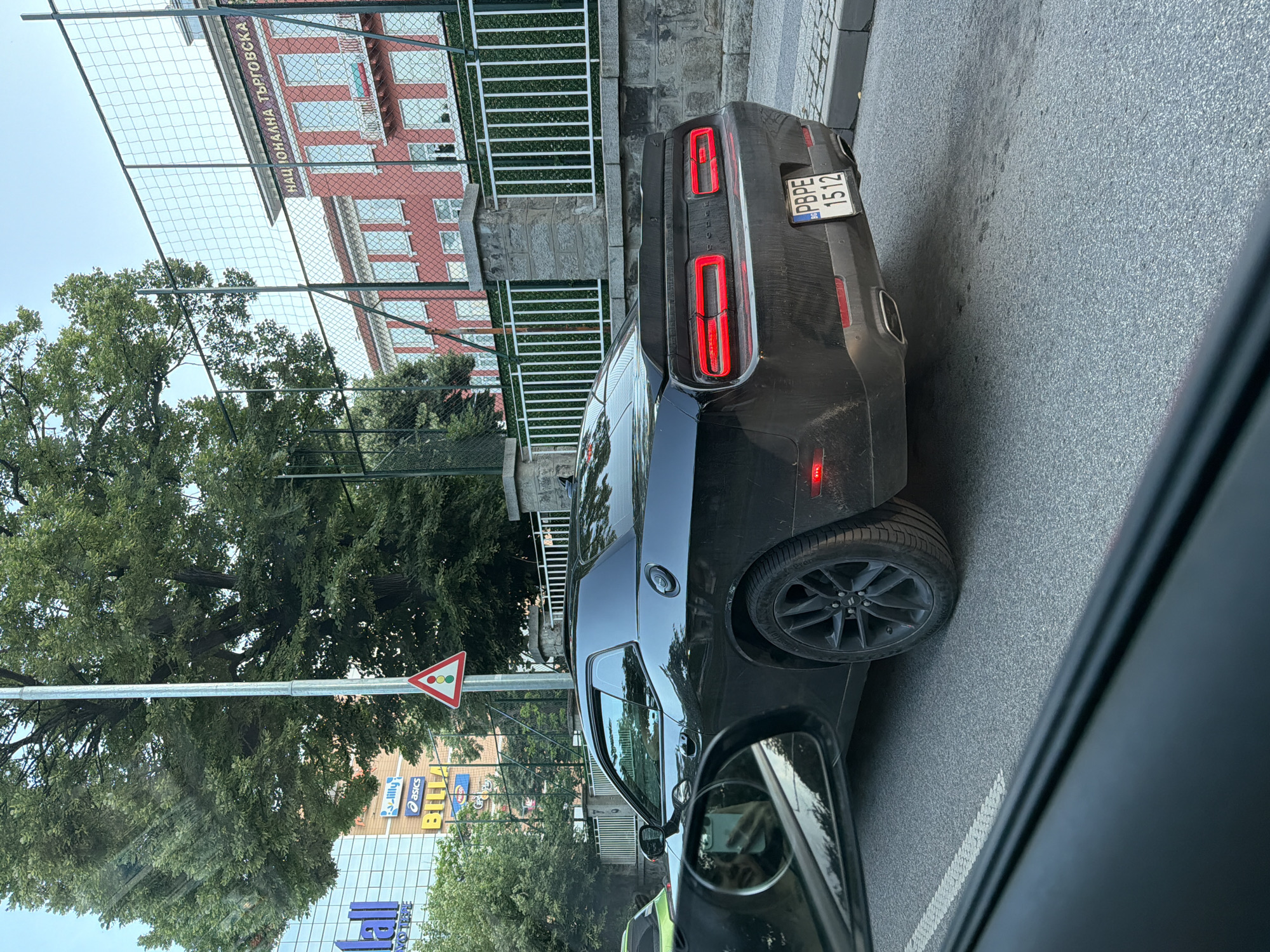 The image captures an urban street scene in Plovdiv, Bulgaria, on a clear day. The photo is taken from inside a vehicle, looking towards the right.
A prominent subject is a dark gray or black Dodge Challenger car, visible from its rear-right side, likely stopped or parked on the asphalt road. Its distinctive rectangular LED taillights glow red. The car features black multi-spoke wheels. A Bulgarian license plate, "PBPE 1512" with a "BG" country identifier, is attached to its rear.
In the background, to the left of the Challenger, stands a red brick building with white-framed windows, partially obscured by a white metal fence and green trees. Cyrillic text, "НАЦИОНАЛНА ТЪРГОВСКА" (National Trade), is visible on the building, indicating a school or commercial establishment.
Further left, beyond more trees, a modern building with a glass facade is visible, displaying commercial signs for "BILLA," "asics," and "lilly." A triangular traffic sign with a red border and three colored circles (red, yellow, green), possibly indicating a traffic light ahead or a give-way junction, is mounted on an overhead pole.
The immediate foreground shows the asphalt road surface and a portion of the side mirror of the vehicle from which the picture was taken. The setting suggests an active city street with a blend of institutional and commercial buildings, alongside natural elements.
