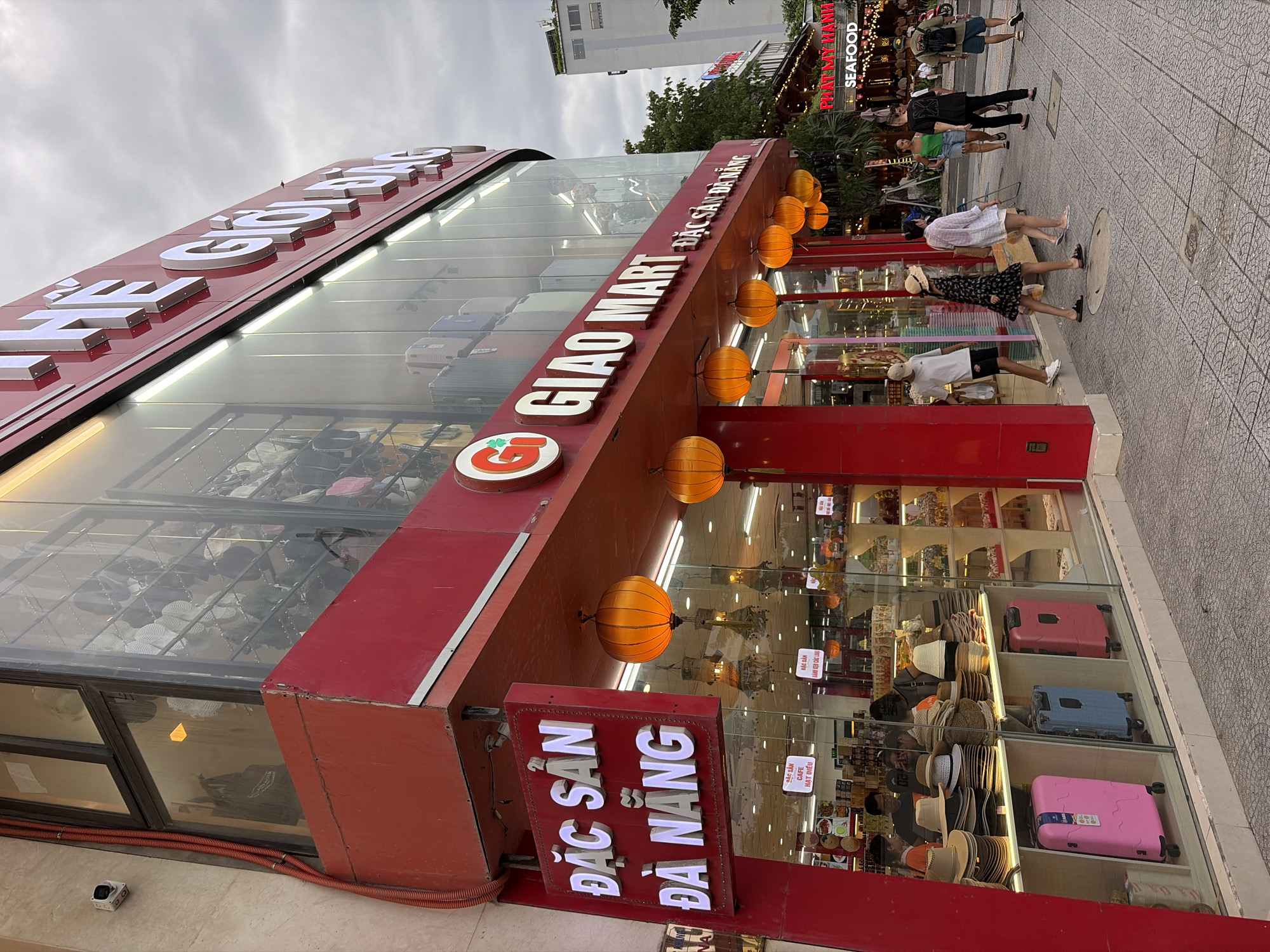 The image shows a storefront in Đà Nẵng, Vietnam. The store is named "GIRO MART" and has a large red facade with white lettering and illuminated signage. Above the entrance, there is a large sign that reads "ĐẶC SẢN ĐÀ NẴNG" (Specialties of Da Nang). Orange lanterns are hanging from the awning of the store. The storefront features large glass windows displaying a variety of goods, including hats, suitcases, and other items. Several people are walking on the sidewalk in front of the store. In the background, there are other buildings and trees. The sky is overcast.