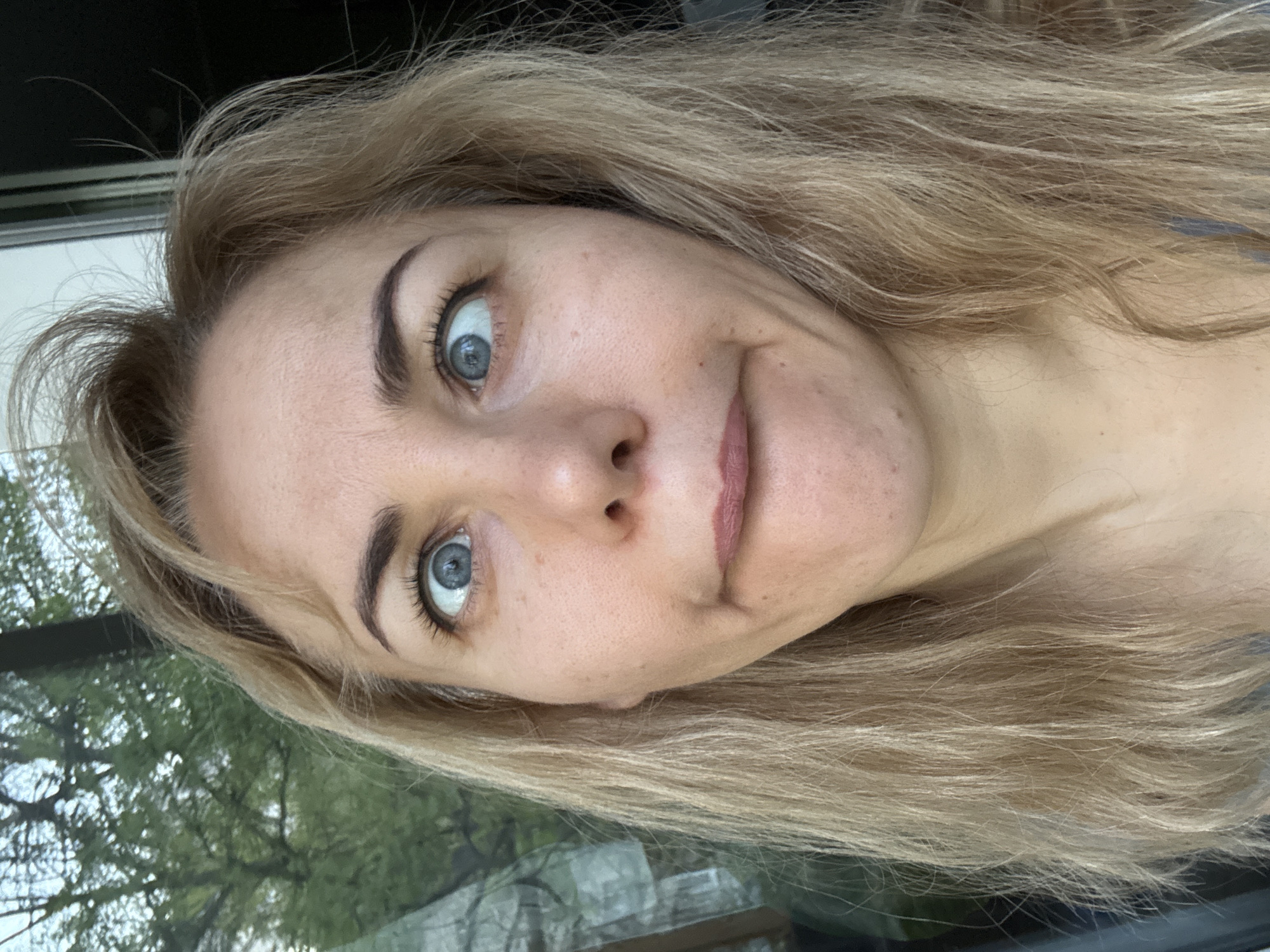 A close-up, tilted selfie shows a blonde woman with fair skin and blue eyes making a slightly quirky facial expression. Her mouth is slightly pursed and angled, and her eyes are wide. Her hair is long and layered, with a mix of light and dark blonde shades. The background shows a window with blurry green trees visible outside, suggesting an indoor setting near a window, possibly a building in an unspecified urban area. No specific city or location is discernible.
