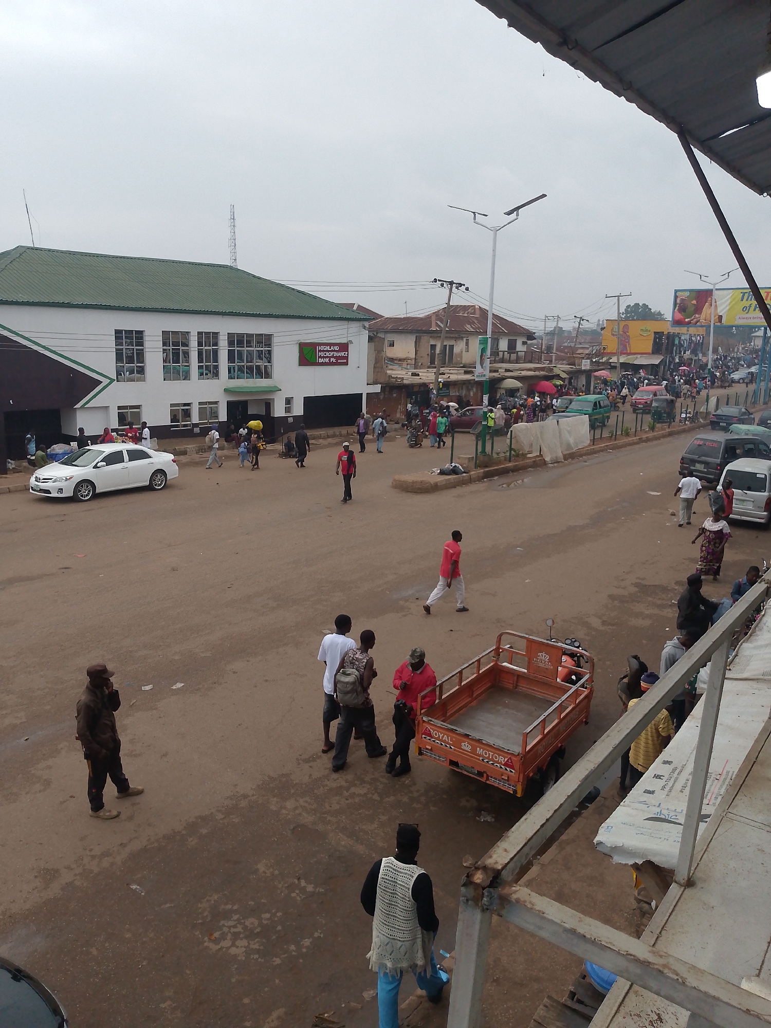 Here is a description of the image:
The image shows a street scene in Bukuru, Nigeria. The view is from an elevated position, possibly a building or an upper floor. The main subject is a wide, unpaved street with people walking, and vehicles moving.
On the left side of the street, a white two-story building is visible with a green roof and a sign for "HIGHLAND MICROFINANCE BANK Plc". Several people are near the building. To the right of the building is a streetlight.
In the foreground are groups of people, and a Royal Motor tricycle with an empty cargo box. The people are of varying ages and dressed in casual clothing. Some appear to be interacting with each other.
The street is somewhat crowded further back, with more people and vehicles. There are also other buildings and structures visible in the distance, with commercial signs along the street. The sky is overcast.
The ground is an earthen color, appearing somewhat dry. The scene depicts a normal day in a moderately busy commercial area in Bukuru, Nigeria.