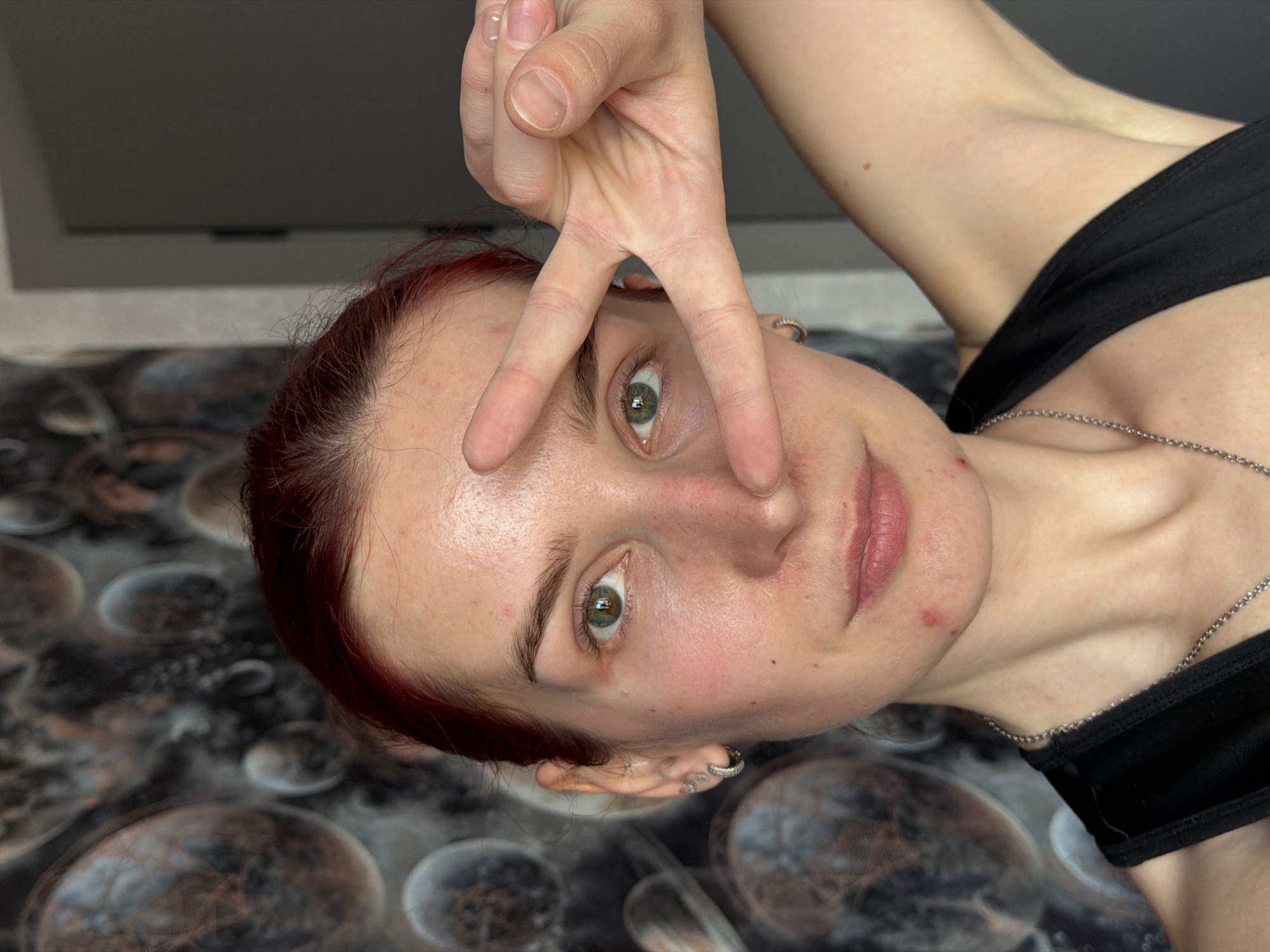A young woman with red hair and fair skin is depicted from a close-up, low-angle perspective. She is wearing a black top and a silver chain necklace. She is making a peace sign with her right hand over her left eye. She has multiple earrings on both ears. The background shows a patterned surface and a wall. There is no evidence to support a location in the city of Chaikovsky, Russia.