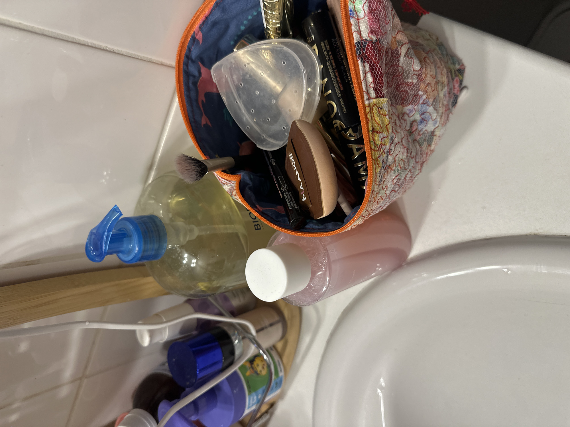 The image shows a collection of toiletries and cosmetics laid out on a white tiled surface, likely a bathroom counter. In the foreground, a clear plastic bottle with a blue pump dispenser contains a yellow liquid, possibly hand soap or lotion. Beside it, a clear pink liquid is in a bottle with a white cap.
A mesh cosmetic bag with an orange zipper is partially open, revealing various makeup items. Among them are a black mascara tube with visible text including "VOLUME MILLION LASH," "MADE IN ITALY," and brand names. There's also a brown makeup sponge and what appears to be a clear plastic cap with perforations. A makeup brush with a dark handle and light bristles is also visible.
Below these items, a white wire basket holds several smaller bottles and containers, some with blue caps. One bottle has a green label with cartoon characters. The overall scene suggests personal grooming or preparation. The setting appears to be indoors, in a bathroom with white tiled walls and a white sink. There are no people visible in the image. The lighting is bright, indicating daytime or good artificial lighting. The text on the mascara tube suggests the products may be of European origin, with references to Paris and Italy.