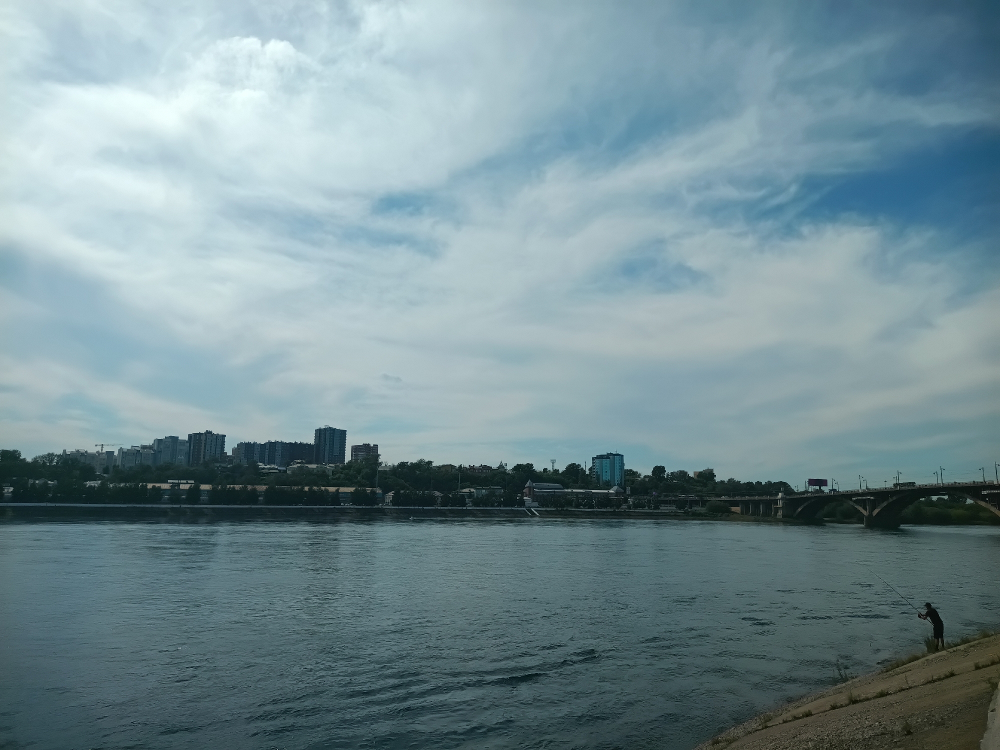 The image captures a wide view of the Angara River in Irkutsk, Russia, under a cloudy sky. On the far bank, a city skyline is visible with several high-rise buildings and trees lining the shore. A large bridge, likely the Irkutsk Railway Bridge or one of its adjacent spans, crosses the river in the distance on the right side of the frame. In the foreground on the right bank, a lone figure is actively fishing from the shore. The river itself is a deep blue, with visible ripples and subtle textures on its surface. The embankment in the foreground is made of concrete and gravel. The overall scene suggests a daytime setting with overcast weather.