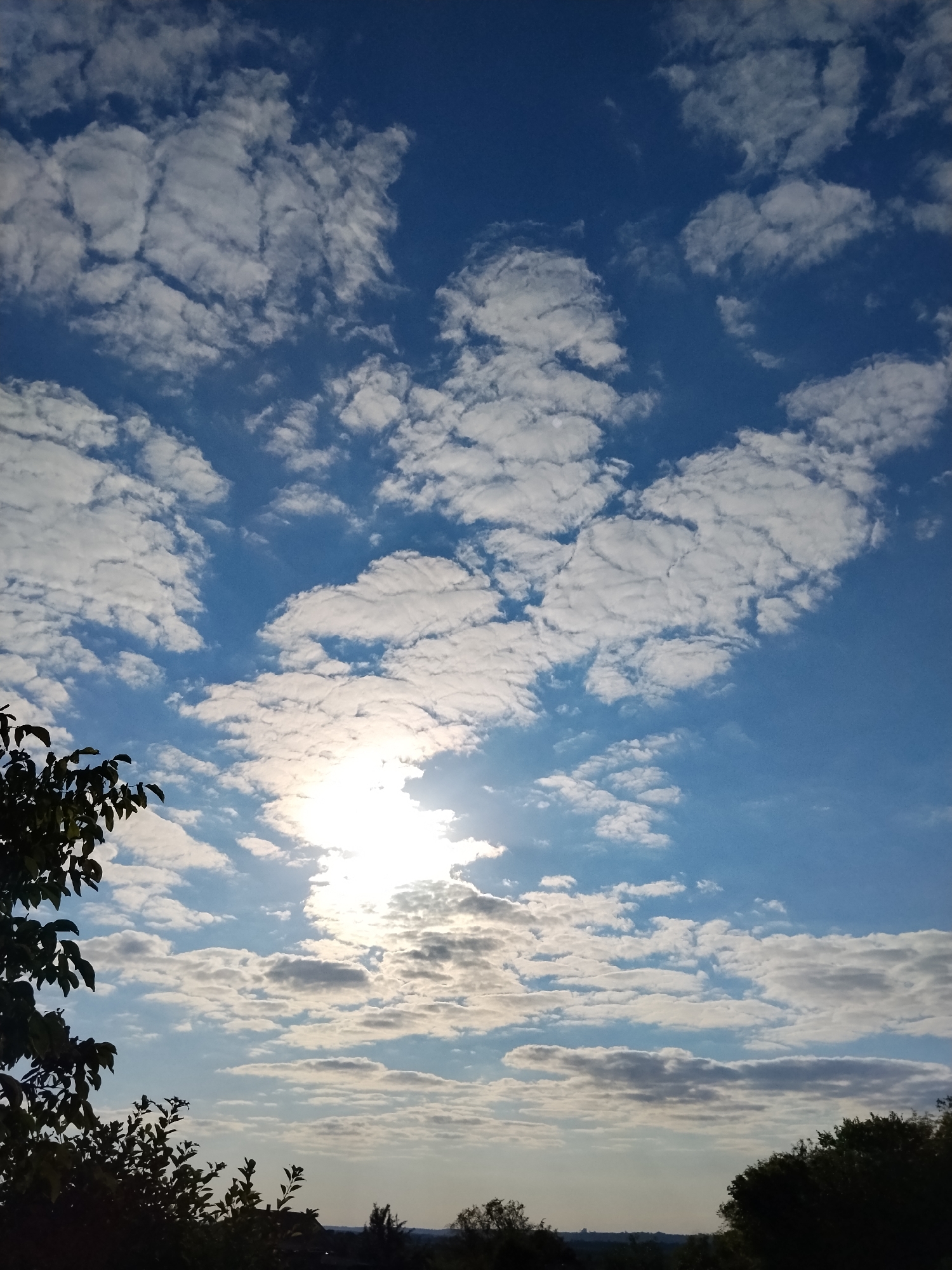 The image depicts a bright, sunlit sky with scattered clouds over a silhouetted landscape. The sun is prominently visible, partially obscured by clouds and positioned slightly off-center towards the left. The sky is a vivid blue, transitioning to a lighter hue near the horizon. The clouds are white and fluffy, with some appearing more wispy and elongated. In the foreground, dark silhouettes of trees and foliage are visible along the bottom edge of the frame. The distant horizon shows subtle hints of structures and terrain, suggesting a rural or semi-rural environment, consistent with a location in Ustyvytsia, Ukraine. The overall scene is serene and indicative of daytime, likely late afternoon or early morning given the sun's angle.