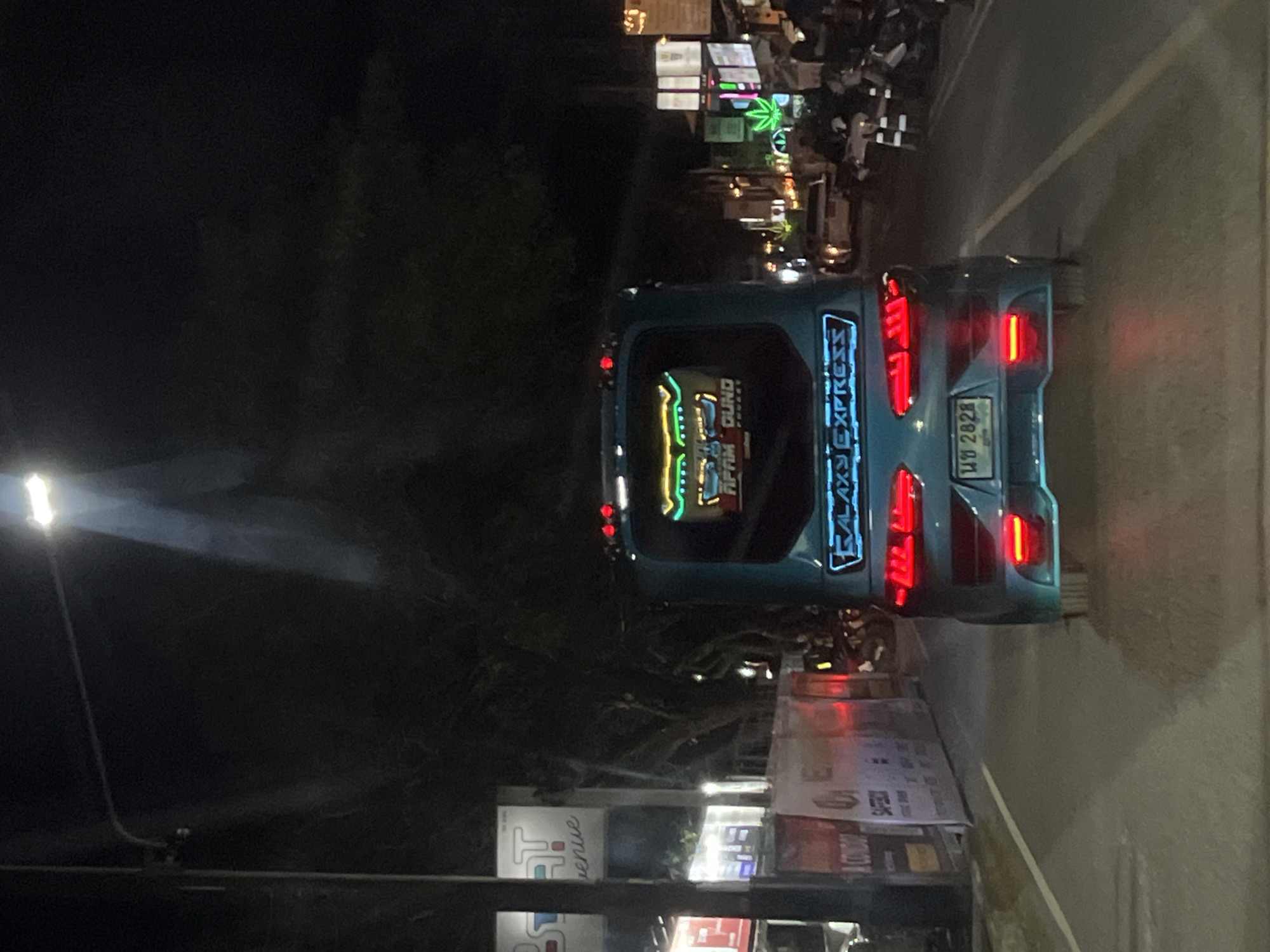 This is a nighttime, outdoor shot of a turquoise bus with neon lights. The bus is the central subject, with its rear facing the viewer. The "GALAXY EXPRESS" is written in neon blue lettering on the back of the bus, and there are red taillights on either side. The license plate is partially visible and appears to have Thai characters and the number "2828". There are colorful neon lights on the back of the bus that spell out "FIFTH DUND". The bus is on a street at night, with a street light visible on the left side of the image. In the background, there are more lights and some trees. Some people are visible in the background, suggesting a bustling environment, possibly a street market or an entertainment area. The overall atmosphere appears lively due to the prominent neon lights.
