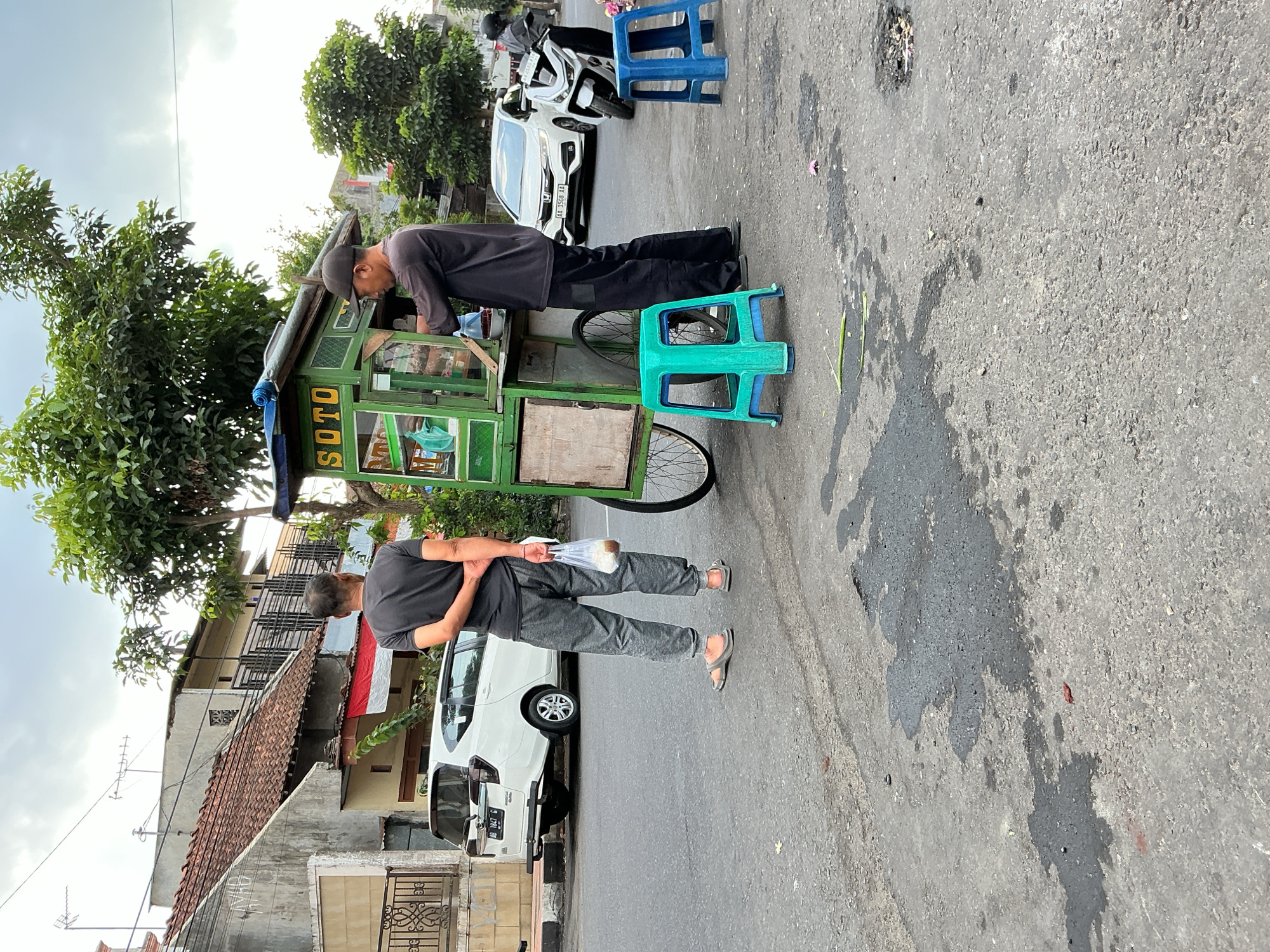 A street vendor is preparing food at a mobile cart labeled "SOTO" in the city of Magelang, Indonesia. The cart is green and mounted on a wheeled frame. A customer stands to the left of the cart, holding a plastic bag with food. The vendor, wearing a dark shirt and cap, is seen behind the cart, engaged in serving food. Two plastic stools, one teal and one blue, are placed in front of the cart. In the background, white cars and a motorcycle are parked on the side of the road, and residential buildings line the street. The sky is overcast.