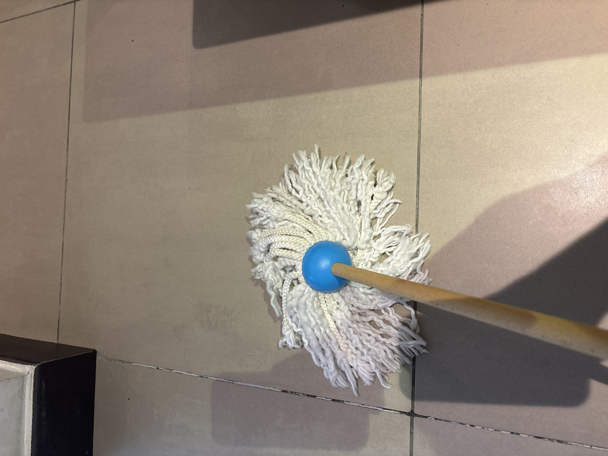 A close-up, eye-level shot depicts a white string mop with a blue spherical head resting on beige tiled flooring. The mop's wooden handle extends diagonally from the bottom left corner upwards. The tiles are arranged in a grid pattern, with visible grout lines. A dark, possibly wooden, structure with a black edge occupies the upper left corner of the frame, casting a shadow on the tiles. The lighting suggests an indoor environment, possibly a home or commercial space. The scene is static, with no discernible action or interaction. No people are present in the image. The context of Dubai, United Arab Emirates, is not evident from the visual information provided in the image.