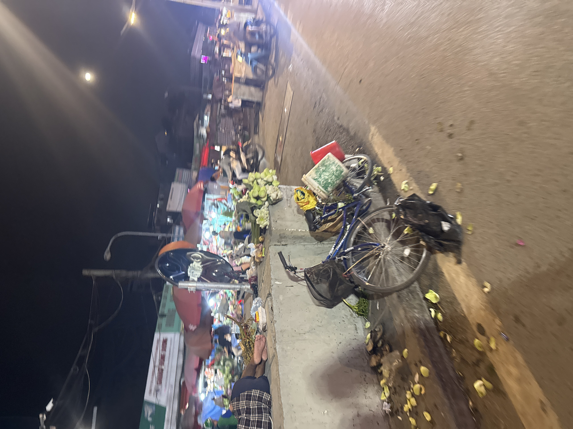The image depicts a street scene at night in Hanoi, Vietnam, likely a market area. A person, clothed in plaid shorts and a dark shirt, is lying down on a concrete ledge, with their feet visible. In the foreground, a bicycle is parked, laden with bags and items, including what appears to be a red bucket and a green container. Scattered around the base of the ledge are fallen leaves or flower petals, along with some debris. The background shows market stalls with red and orange awnings, lit by artificial lights. Several motorcycles are present on the street, with some people riding them, suggesting ongoing activity. The overall impression is one of a bustling, yet somewhat chaotic, nighttime market environment.