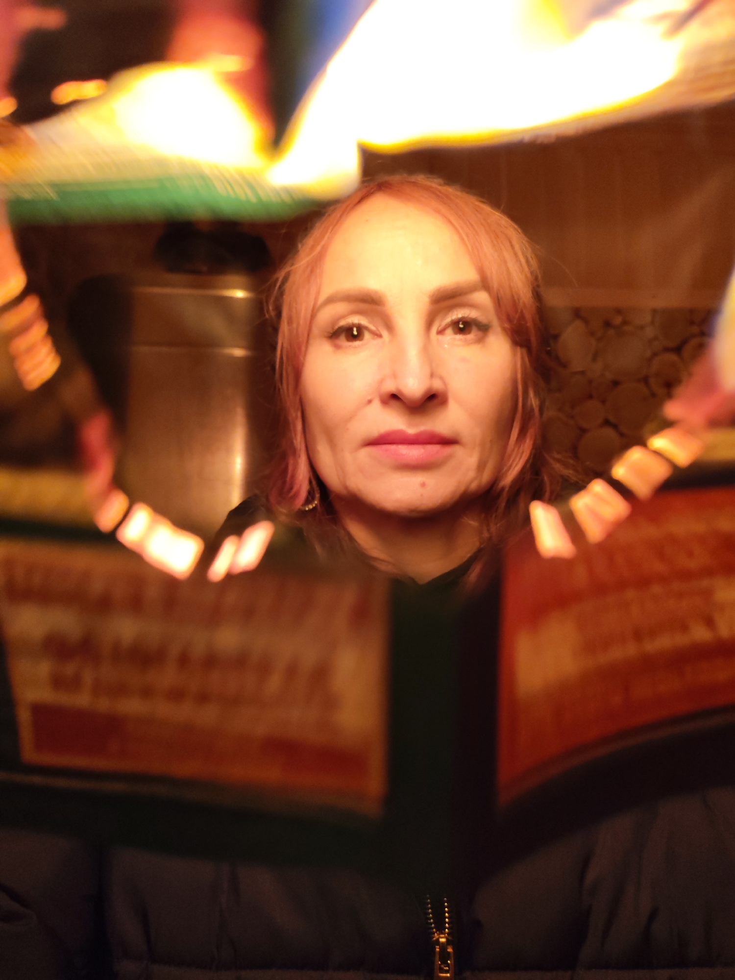 A woman with fair skin and reddish-blonde hair looks directly at the camera. She has light eyes, prominent cheekbones, and is wearing lipstick. She is framed by an out-of-focus, angular shape of orange and yellow, likely a book or pamphlet held up close to the lens. She is wearing a dark-colored top or jacket, the zipper of which is visible at the bottom of the frame. The background behind her appears to be an interior setting, including what looks like a metal cabinet or appliance, and a wall possibly decorated with round, wooden accents. The location is unknown.