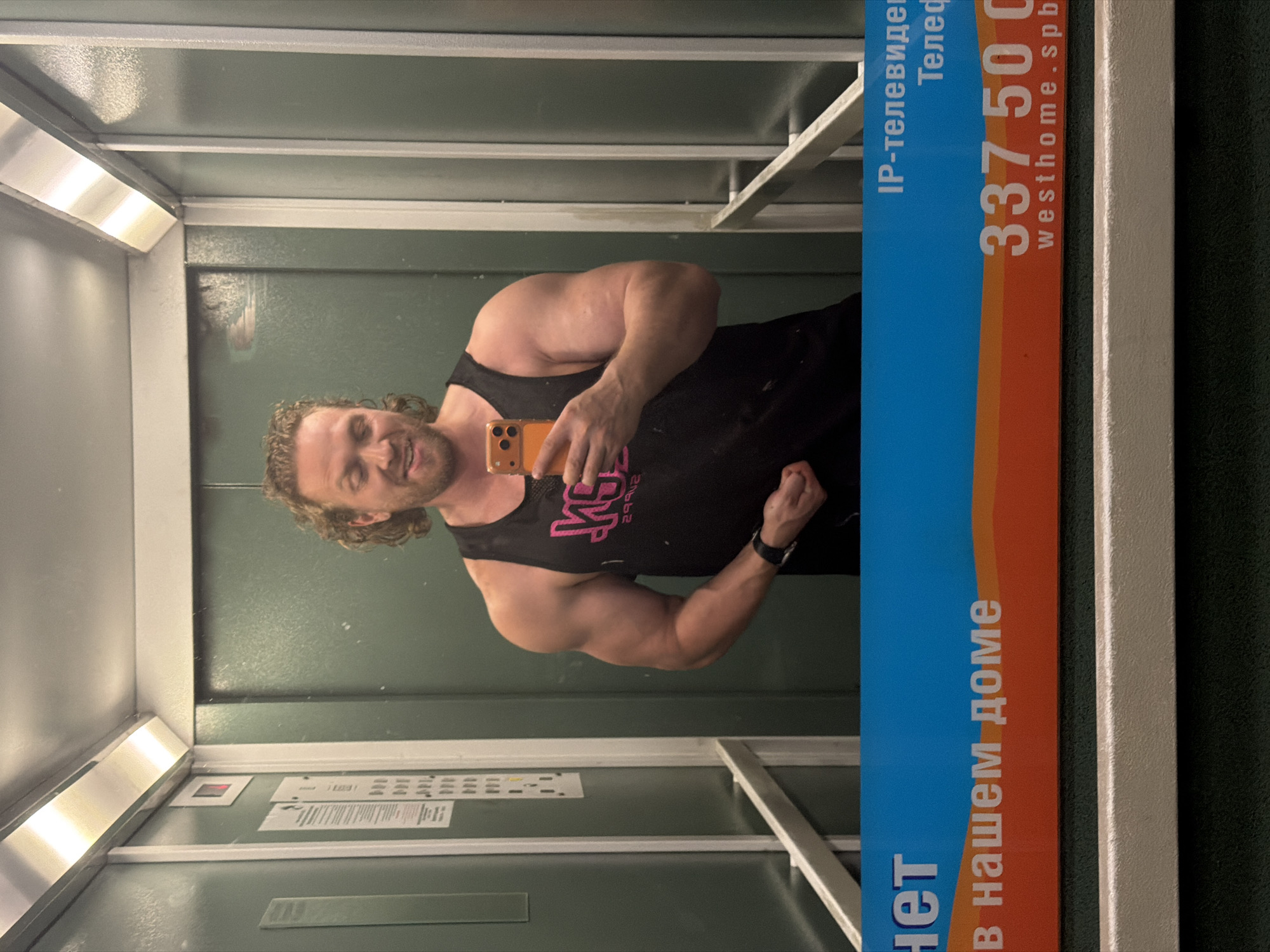 A muscular, fair-skinned man with blonde, curly hair and a beard poses for a mirror selfie in an elevator. He is wearing a black tank top with a pink logo and an orange phone in his right hand. The elevator has green walls, silver trim, and bright overhead lighting. A sign with Russian text and a phone number is visible at the bottom of the image, suggesting the location is in Saint Petersburg, Russia.