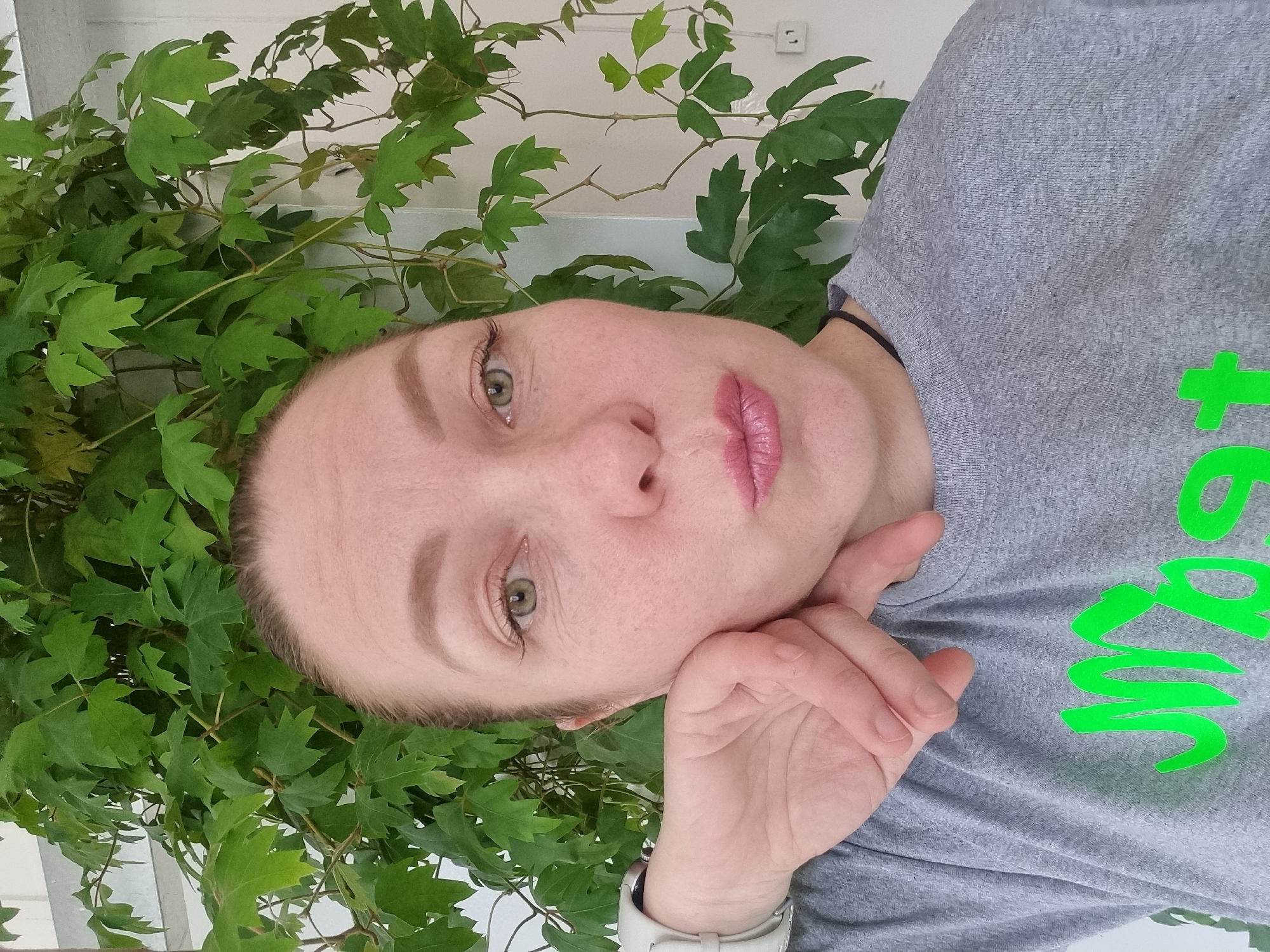 A woman is posing for a selfie in front of a green plant. The woman has fair skin, light brown hair pulled back, and is wearing a gray t-shirt. Her left hand is positioned near her chin. A white smartwatch is on her wrist. Behind the woman, there is a plant with green leaves. A white wall is partially visible in the background behind the plant. It is daytime, and the location is unknown.