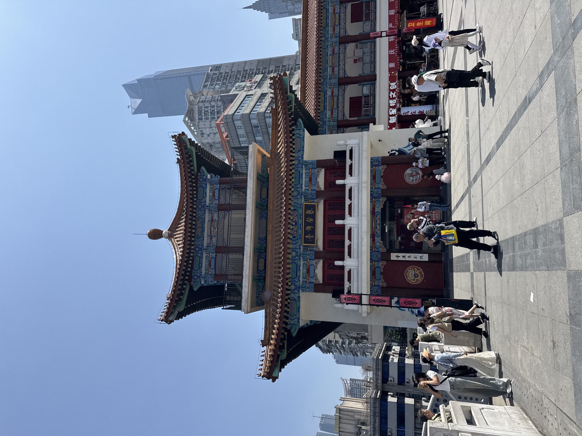 The image captures a traditional Chinese structure against a backdrop of modern buildings in the city of Yuzhong District, China. The structure is a multi-tiered building with a red and blue ornate roof, featuring curved eaves and intricate designs. There are Chinese characters inscribed on the building.
The foreground shows a public square with people. Some people are walking, others are standing, and some are looking at the structure.
Behind the traditional structure, modern high-rise buildings are visible, creating a contrast between old and new architecture. The sky is clear and blue.