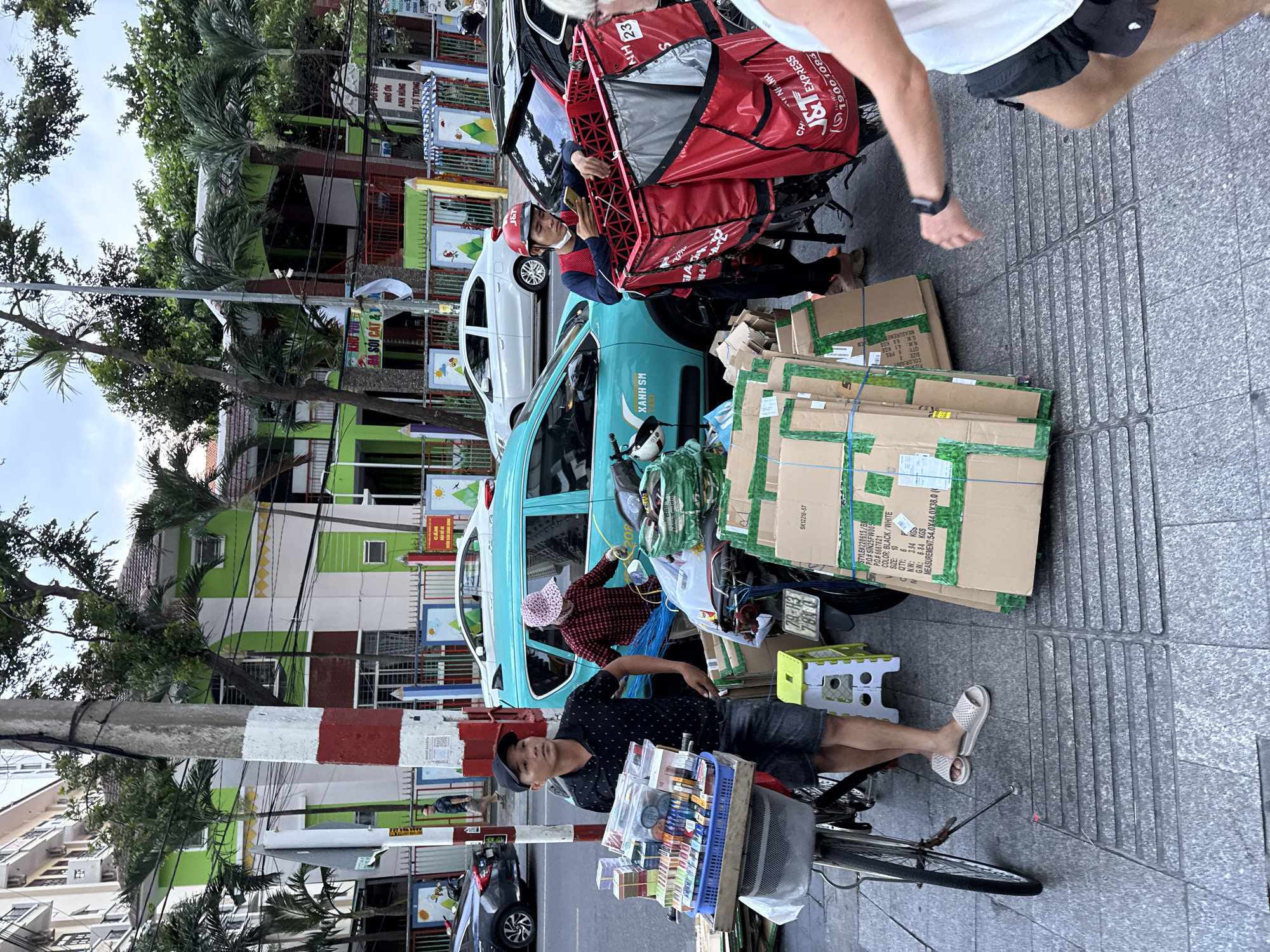 In Nha Trang Ward, Vietnam, on a street, a man wearing a black polo shirt and denim shorts stands beside a bicycle loaded with various items, including boxes and what appear to be cigarettes or small packaged goods. To his right, another person, a delivery person wearing a red vest and helmet, is looking at their phone while leaning against a red delivery cart. The cart is attached to a motorcycle. In the background, a turquoise vehicle is parked, and several buildings are visible, one with a distinctive green and white facade. The street is paved with textured tiles, and there are palm trees and other vegetation adding to the urban environment. The scene depicts a street vendor and a delivery person operating in a busy urban setting.