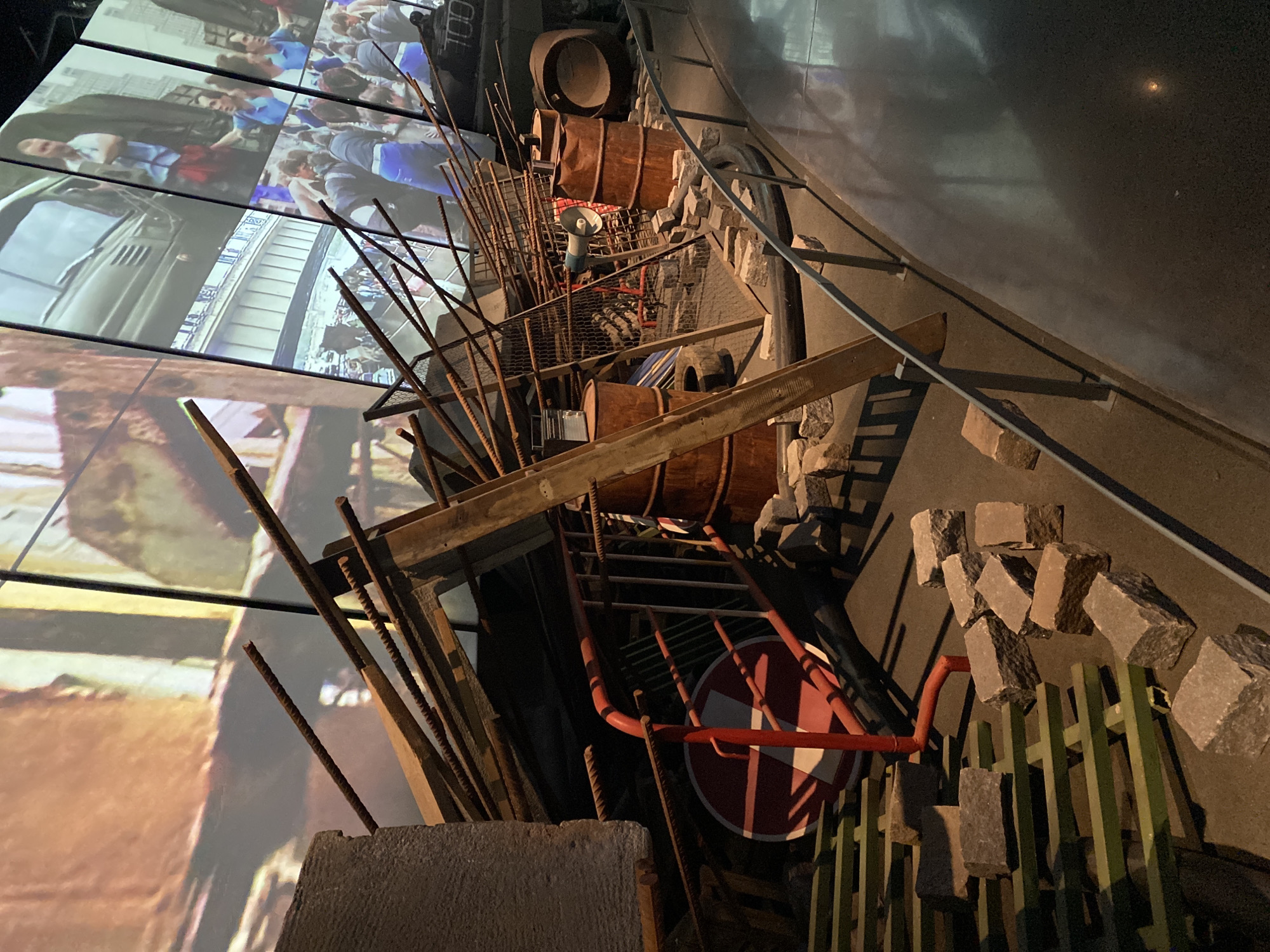 The image shows a barricade exhibit at the Boris Yeltsin Presidential Center in Yekaterinburg, Russia. The barricade is constructed of various objects, including wooden barrels, metal rods, a street sign with a red and white diagonal stripe, pieces of wood, concrete blocks, and cobblestones, all piled together. In the background, a large screen displays footage of people, vehicles, and city streets, likely depicting scenes of civil unrest or protests. The exhibit is situated indoors, with a dark wall and a walkway visible to the side. The lighting is dim, creating a dramatic effect. The purpose of the exhibit appears to be to recreate or simulate a barricade that might be erected during a period of social or political upheaval.