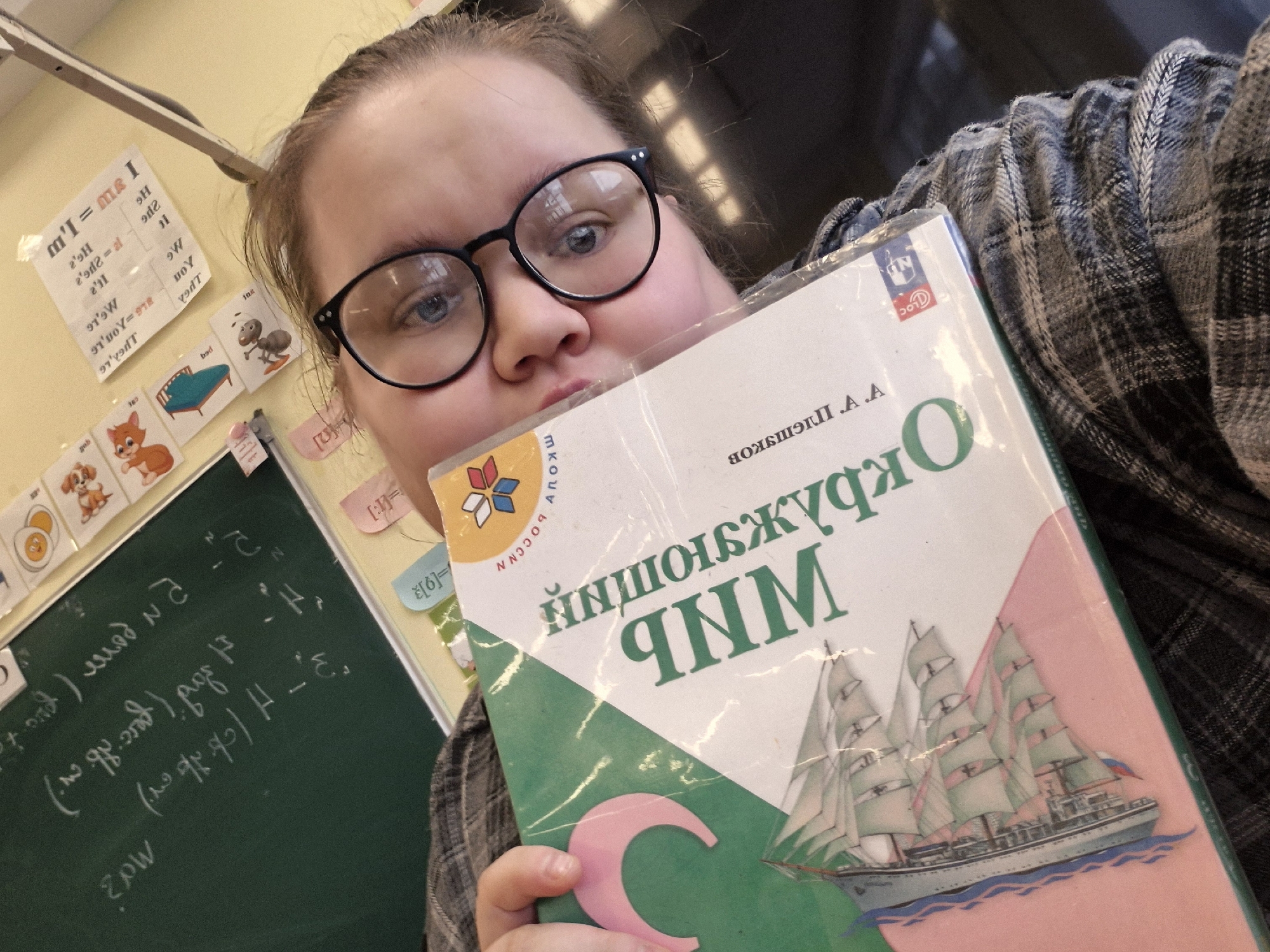 A young woman wearing glasses is holding a Russian language textbook with a ship illustration on the cover. She appears to be in a classroom, as evidenced by the blackboard in the background with mathematical equations written on it, and charts with words or information on the wall. The textbook seems to be titled "Okruzhayuschiy Mir" (Окружающий мир), which translates to "Surrounding World." The textbook has a Russian flag logo. The classroom setting and textbook suggest this is in a school environment. There is no information available in the image to indicate this location is in the city of Chaikovsky, Russia.