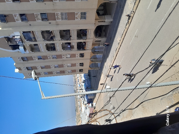A sideways view of a sunlit urban landscape shows a tall building with multiple floors, balconies, and arched openings alongside a wide road. Several people are walking on the road and along the edge of the building. Shadows from overhead power lines and a street light fall across the road. In the distance, more buildings stretch out under a clear blue sky.