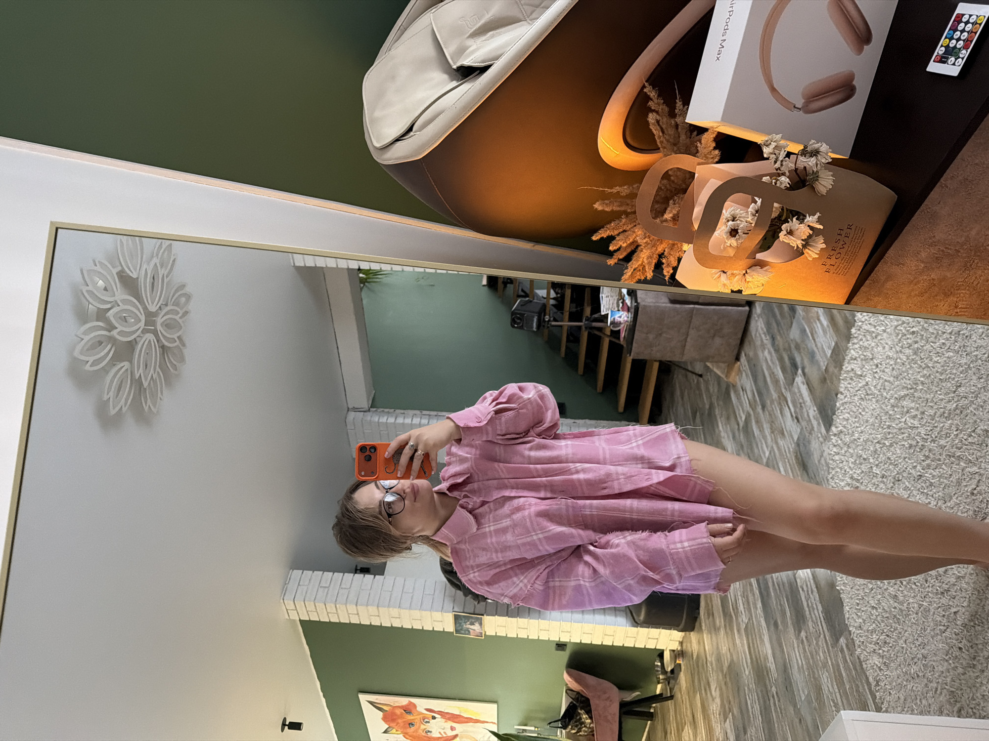 A young woman takes a mirror selfie in a brightly lit room in Большая Шильна, Russia. She has shoulder-length brown hair and wears glasses, a pink plaid oversized button-up shirt, and what appears to be light-colored shorts or briefs. She holds an orange-cased smartphone in her right hand, positioned to capture her reflection. Several rings are visible on her fingers.
The room features a contemporary design with two-toned walls, one section painted a dark green and another a light neutral color. There is also a partial white brick wall reflected in the mirror. The flooring consists of light wood planks transitioning to a light, shaggy carpet.
Behind the woman, reflected in the mirror, is a modern home interior. A large beige and brown armchair or massage chair with a "G" symbol on it is visible, along with a dark side table. On the table are a pair of rose gold Apple AirPods Max headphones in their box, which clearly displays "Airpods Max" text. Beside them is a brown paper bag filled with dried white flowers, featuring the text "FRESH FLOWER," and a remote control with multiple colored buttons. Dried pampas grass or similar floral arrangements are also placed on the table.
Further into the background, stairs can be seen, and a projector sits on a surface. On the wall behind the woman's reflected figure, there is a framed artwork depicting a fox's head. Another white, intricate floral-patterned wall decoration is mounted higher on the wall. A small black security camera or sensor is installed near the floor on a light-colored wall. The overall ambiance suggests a comfortable and decorated living space.