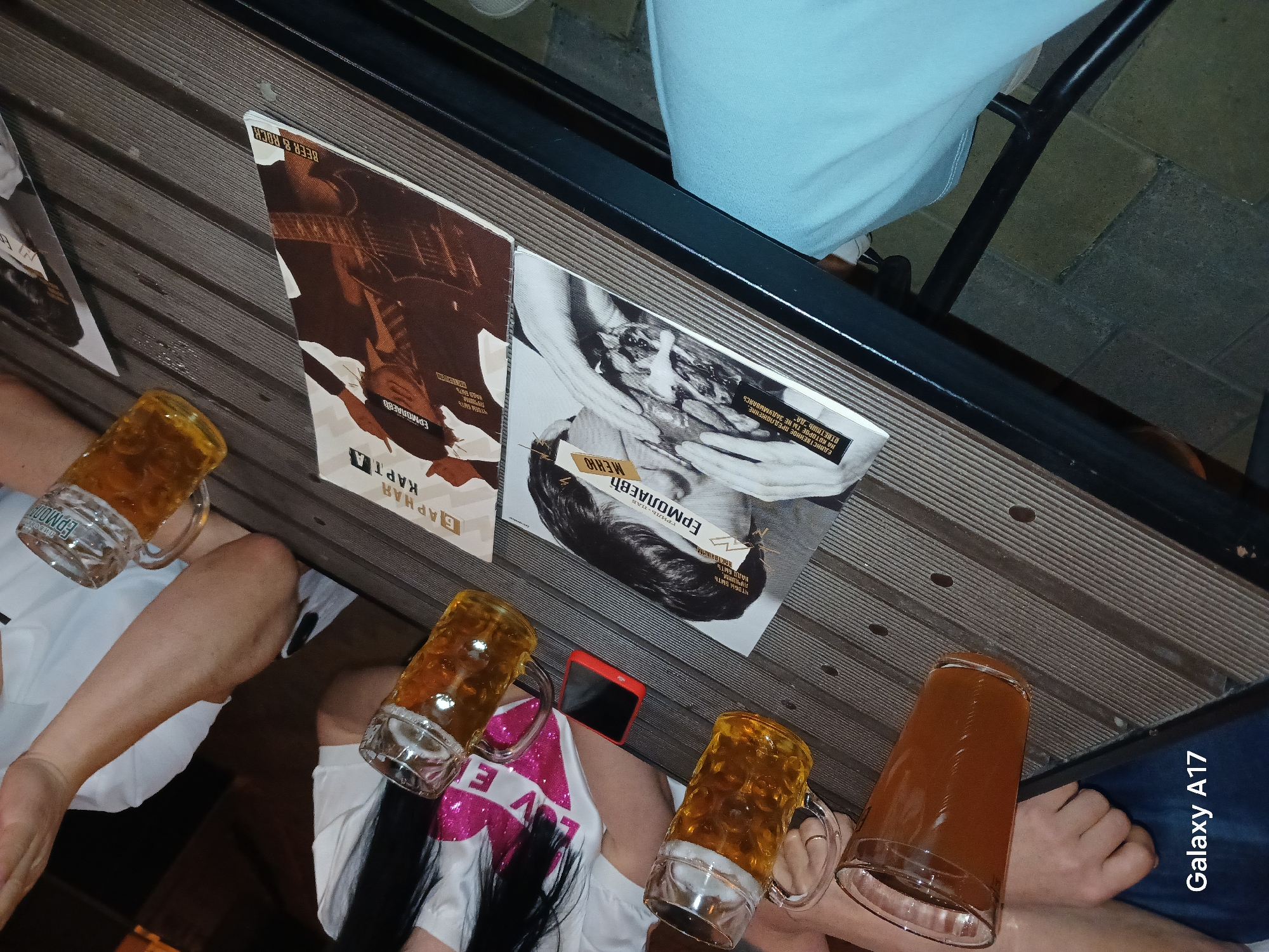 The image, taken in Yekaterinburg, Russia, captures a casual scene at what appears to be an outdoor bar or restaurant. Four beer mugs, filled with what is likely beer, are on a wooden table. Two paper menus for "Ermolaev," a bar and restaurant, are also on the table. The menus have graphics and text in Russian. The "Galaxy A17" branding is visible, indicating the camera used. The scene shows people, partially visible, sitting at the table, with their arms and hands near the beers.