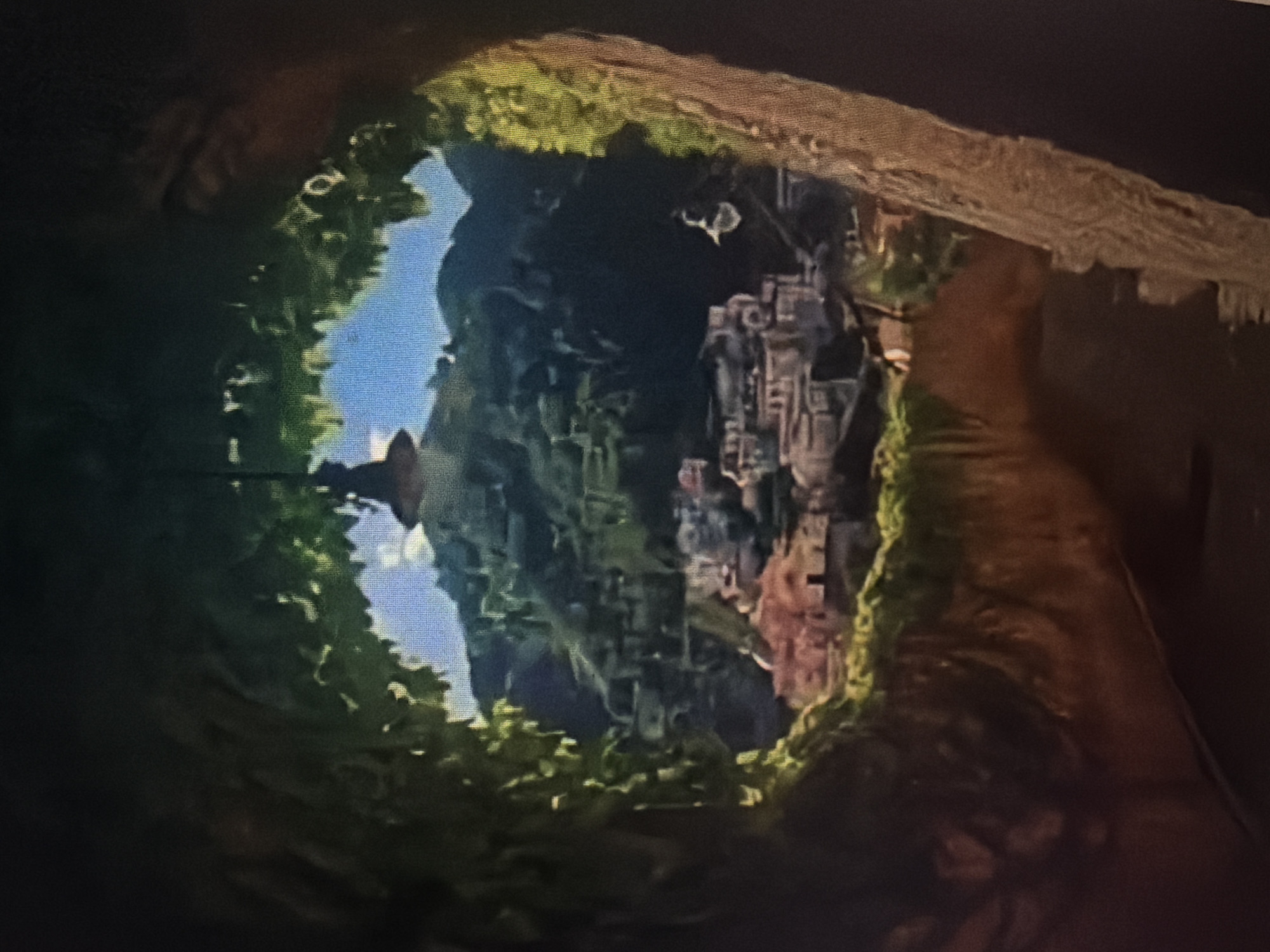The image, taken from what appears to be a rustic vantage point framed by greenery and rock formations, looks out onto a distant village nestled in a valley. The village consists of numerous buildings with terracotta roofs, clinging to the side of a lush, green mountain. The sky is a bright, clear blue, and the overall impression is one of a picturesque, possibly Mediterranean or Southeast Asian, rural landscape. There are no discernible people or specific actions depicted in the scene.
It is not possible to determine the exact location within the city of Baan Mae Nam, Thailand based on this image alone.
