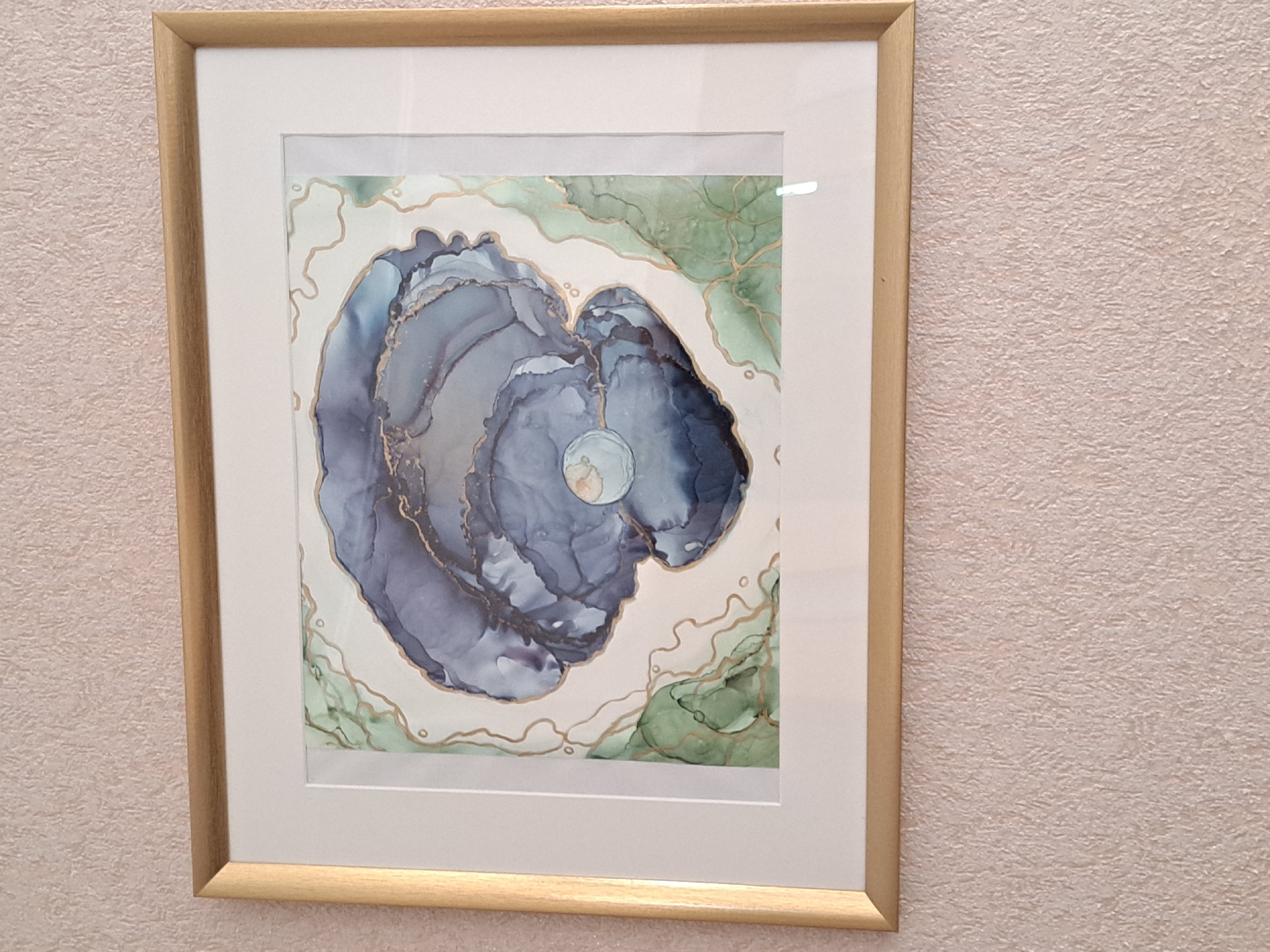 A framed artwork hangs on a textured, light-colored wall. The frame is light wood, and there is a white matte boarder inside of it. The art appears to be an abstract design with blue, green, and gold colors. The central form is a swirling blue shape. The background surrounding the blue form is white with touches of green and gold lines. The location is not discernible from the image.