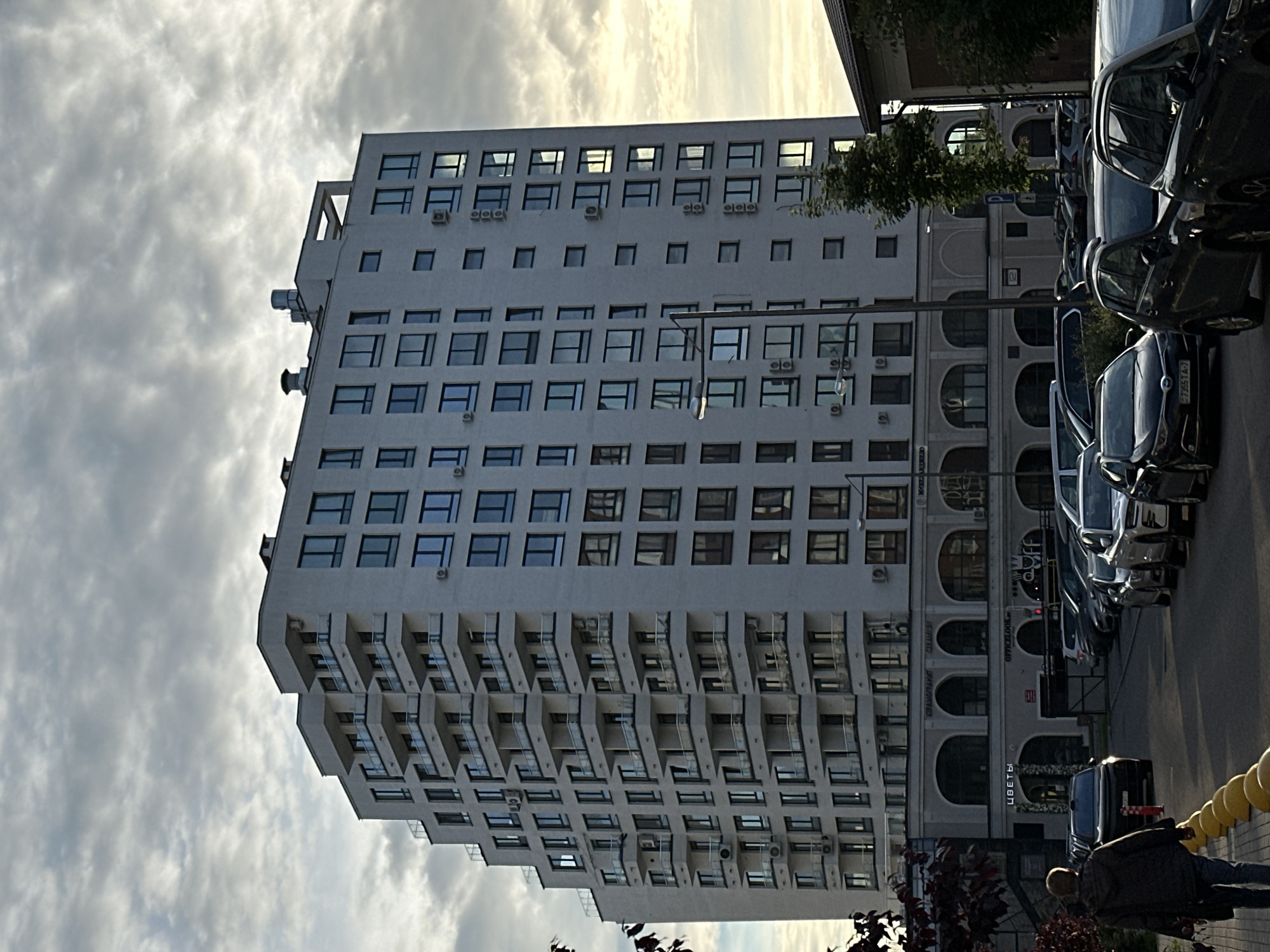An exterior, low-angle shot depicts a modern, multi-story building with many windows and balconies in an urban setting under a cloudy sky. The building is white and appears to be residential or office space, with a row of arched storefronts at its base. In the foreground, a person walks away from the camera on a sidewalk. To the right of the building, a tree and the roof of another structure are visible. Cars are parked in a lot or on the street in front of the building, with a row of yellow bollards along the edge of the sidewalk. The scene appears to be illuminated by natural light, suggesting it is daytime.