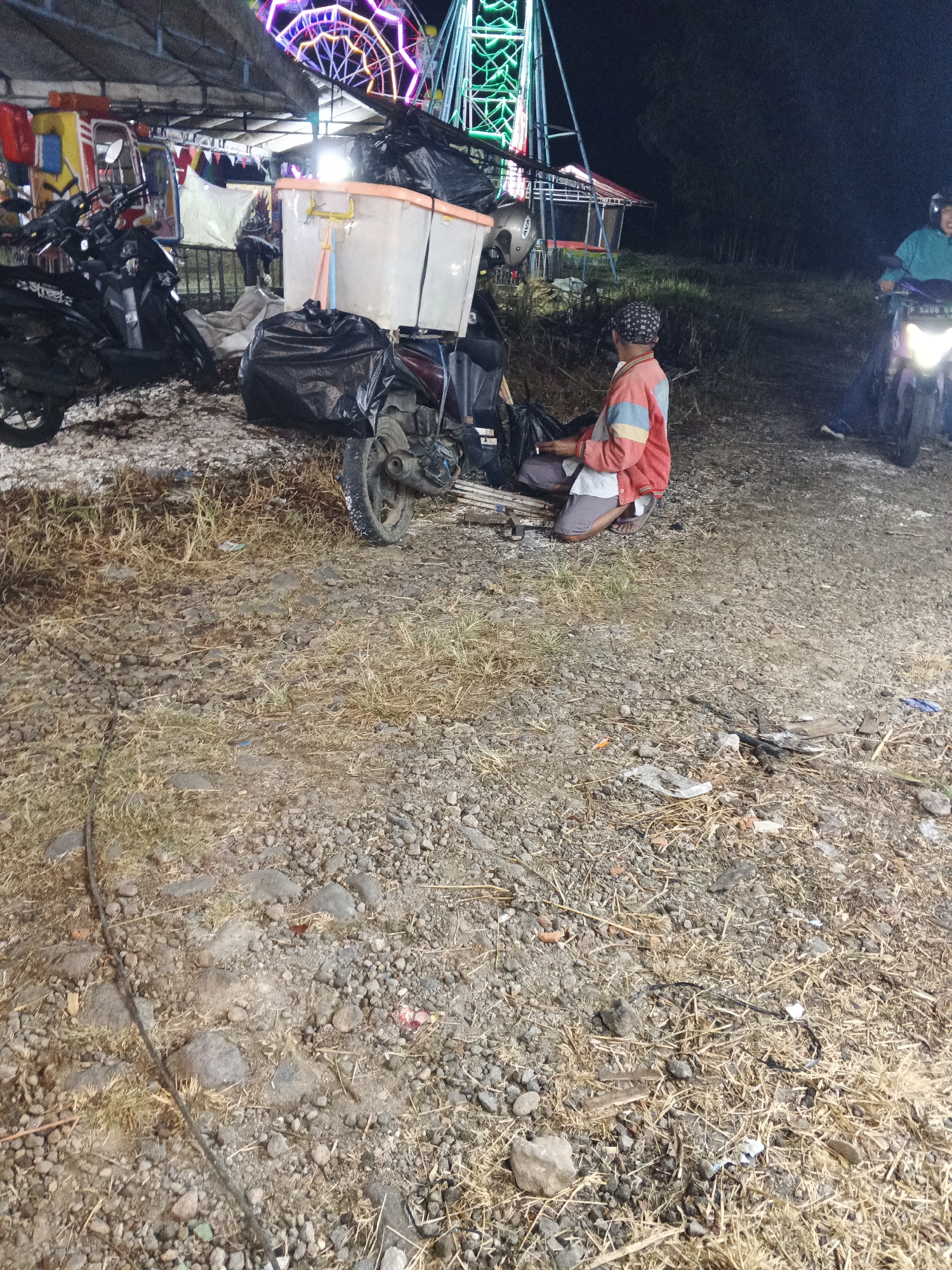 The image depicts an outdoor scene at night, likely a fairground or market area, possibly in Sukabumi, Indonesia, given the context. In the foreground, a person is crouched down next to a motorcycle, which is laden with a large black plastic bag and a translucent plastic container with an orange lid. The motorcycle is parked on a rough, gravelly surface with sparse dry grass. Behind the motorcycle, a small, colorful amusement ride, possibly a carousel or a similar attraction, is visible, illuminated by bright lights in various colors, including pink, purple, and green. Another motorcycle, ridden by a person wearing a helmet, is seen in the mid-ground on the right, approaching from the background. The overall lighting suggests it is after dark, with artificial lights from the amusement ride and the motorcycle headlights illuminating the scene. The environment appears somewhat unkempt, with debris scattered on the ground.