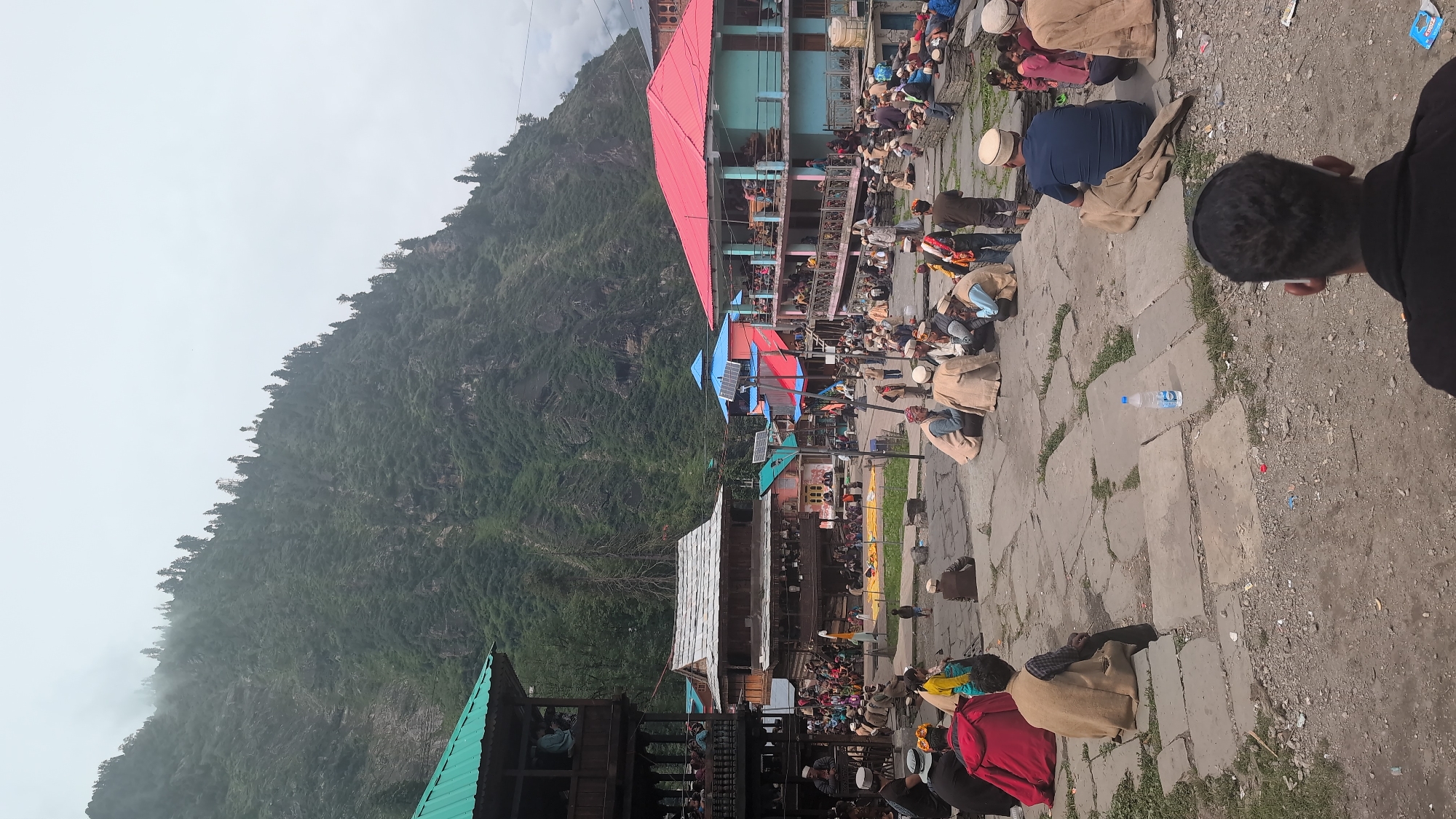 The image captures a lively scene in the village of Malana, India, nestled amidst a dramatic mountain landscape. The architecture is distinctly traditional, with wooden structures featuring multi-tiered balconies and sloping roofs, some adorned with vibrant red and blue hues. People, mostly men, are gathered in the central open area, dressed in traditional attire, many wearing distinctive woolen caps. They are seen sitting, conversing, and observing. Some are seated on the stone paving, while others occupy raised platforms. In the background, a steep, verdant mountain slope rises, partially shrouded in mist, indicating a high-altitude location. The atmosphere appears to be that of a community gathering or a local event. The ground is paved with large, irregular stone slabs, with patches of dirt and sparse vegetation. Various everyday objects are visible, including a water bottle placed on the ground and some scattered debris. The overall impression is one of a cultural snapshot of life in a remote Indian mountain village.