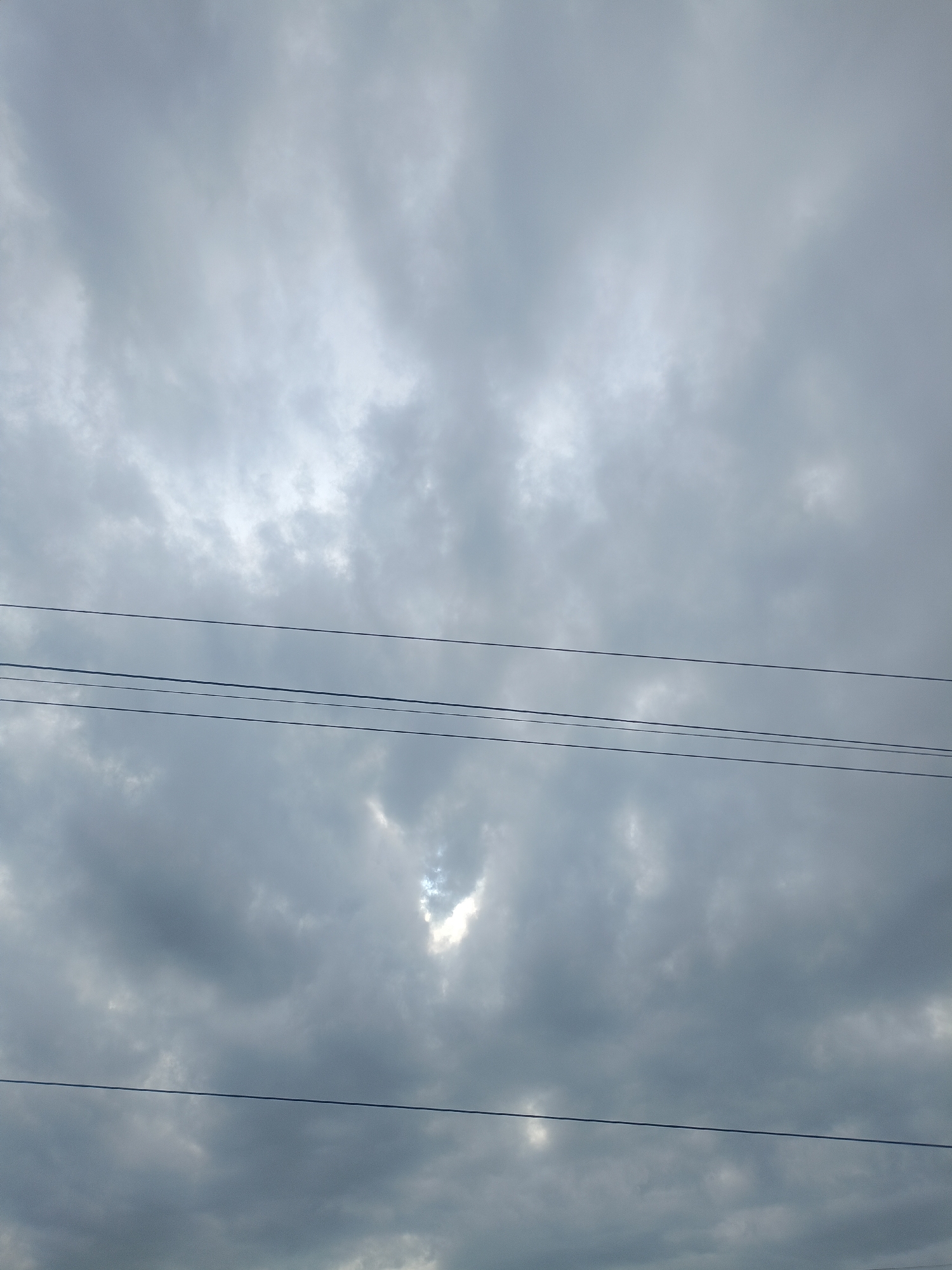 The image shows a cloudy sky in Bireuen, Indonesia. Power lines are visible in the foreground against the backdrop of predominantly grey clouds. A small patch of bright sky is visible between some of the clouds. The overall scene suggests overcast conditions.