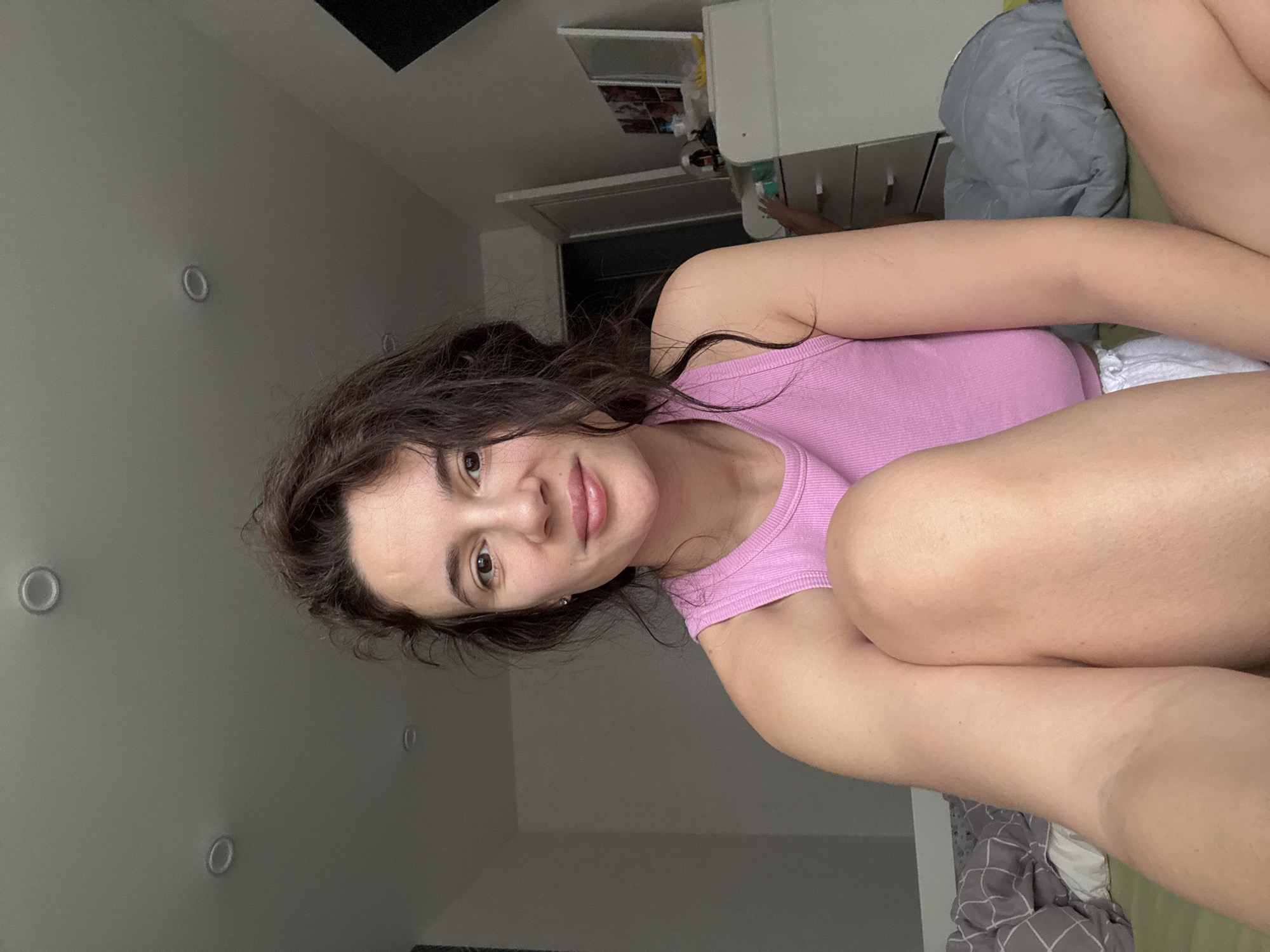 A woman with dark curly hair is lying on a bed. She is wearing a pink tank top. Her legs are drawn up towards her chest. The background includes a white wall with several recessed lights, a white cabinet, and bedding. The location cannot be determined to be Chaikovsky, Russia.