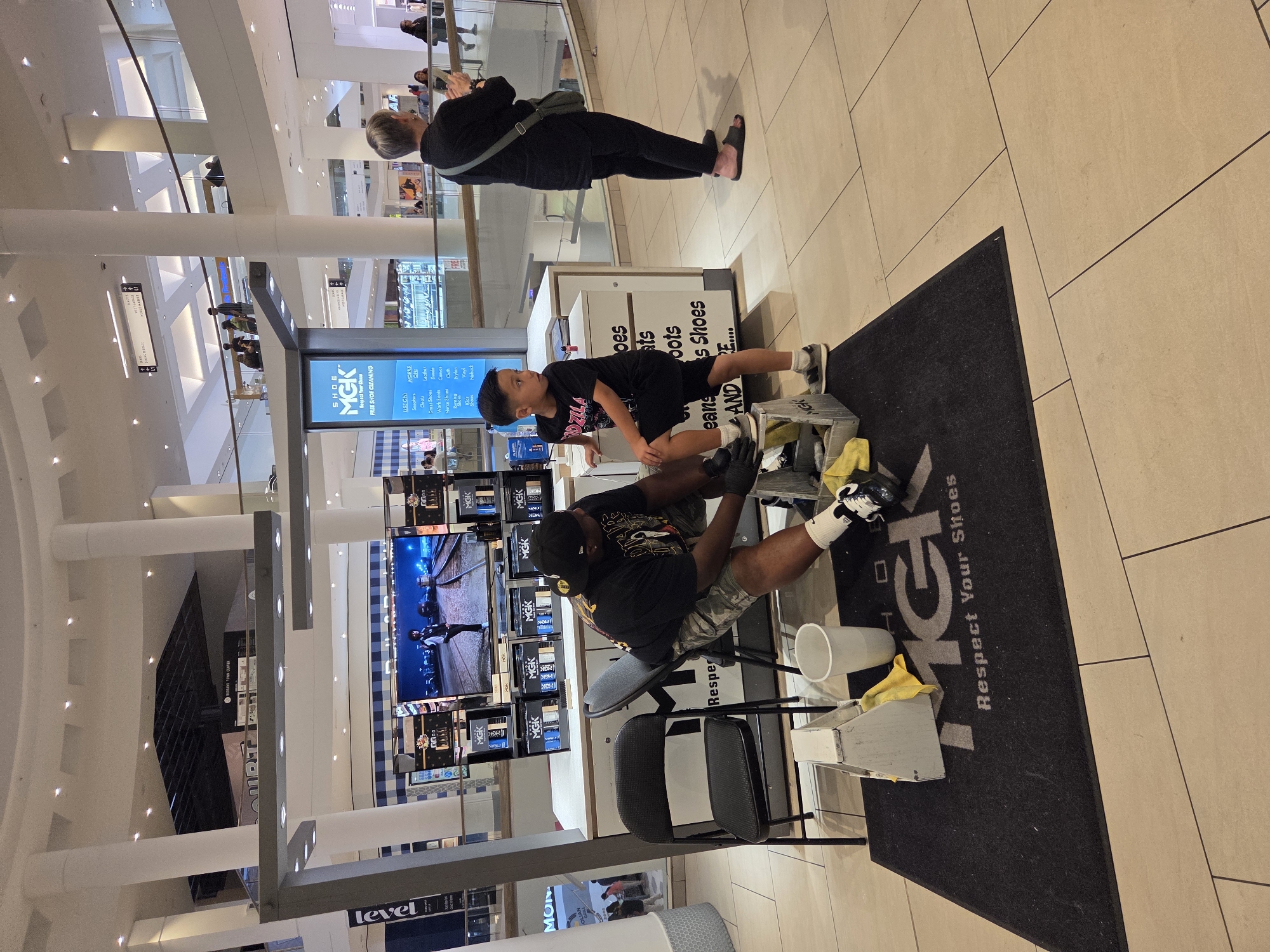 A shoe cleaning service stand is located in a shopping mall, likely in Burbank, California, given the mall's signage. A man wearing a black t-shirt with graphics, camouflage shorts, a black baseball cap, and white socks with black sneakers is seated on a black folding chair, cleaning a shoe. A young boy in a black t-shirt with "GODZILLA" printed on it and black shorts stands next to the man, looking to his left. In front of them, on a black mat with "MGK Respect Your Shoes" printed in white, is a white toolbox containing yellow cloths, and a white plastic cup. Behind the man and boy, a counter displays various shoe cleaning products and a large screen showing an advertisement. Several other shoppers are visible in the background, walking through the mall. The environment appears to be well-lit with ambient mall lighting and fluorescent lights.