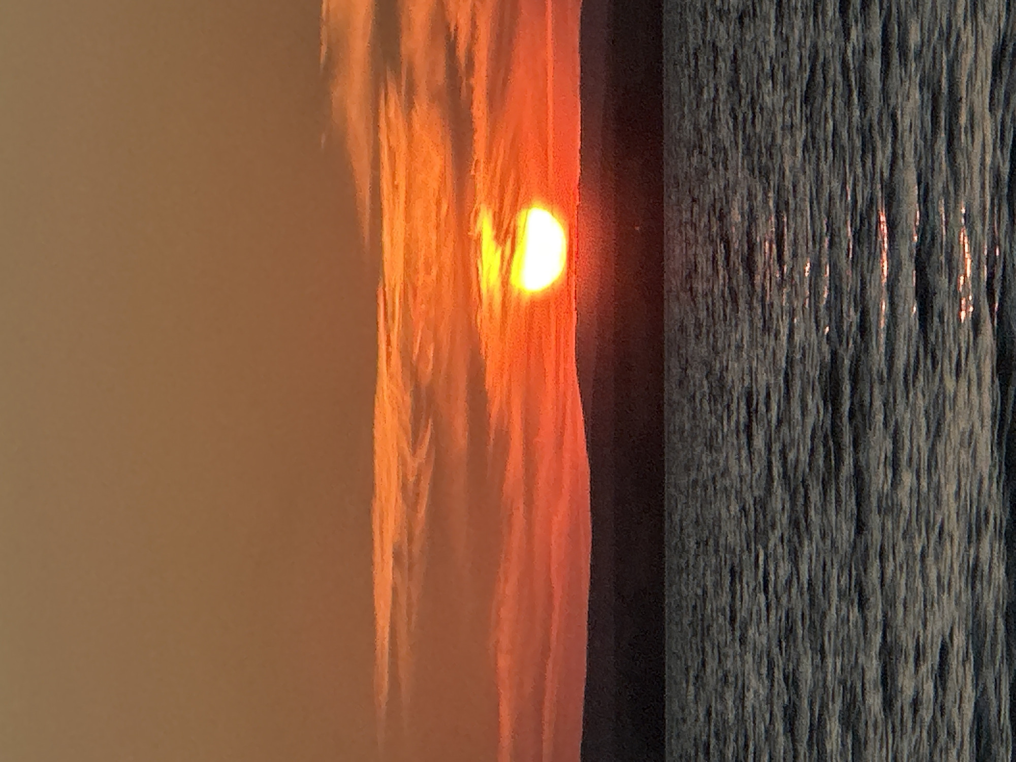 This is a photograph of a sunset over a body of water, possibly the Energetik Reservoir, near the city of Energetik, Russia. The sun, a bright yellow orb with a white center, is low on the horizon, partially obscured by clouds. The sky is a gradient of orange and red hues, with wispy clouds streaked across it. A dark, silhouetted landmass, likely a distant shore or hills, lies beneath the horizon line. The water in the foreground is dark and choppy, reflecting the colors of the sunset. The image is characterized by warm, vibrant colors and a serene, natural landscape.