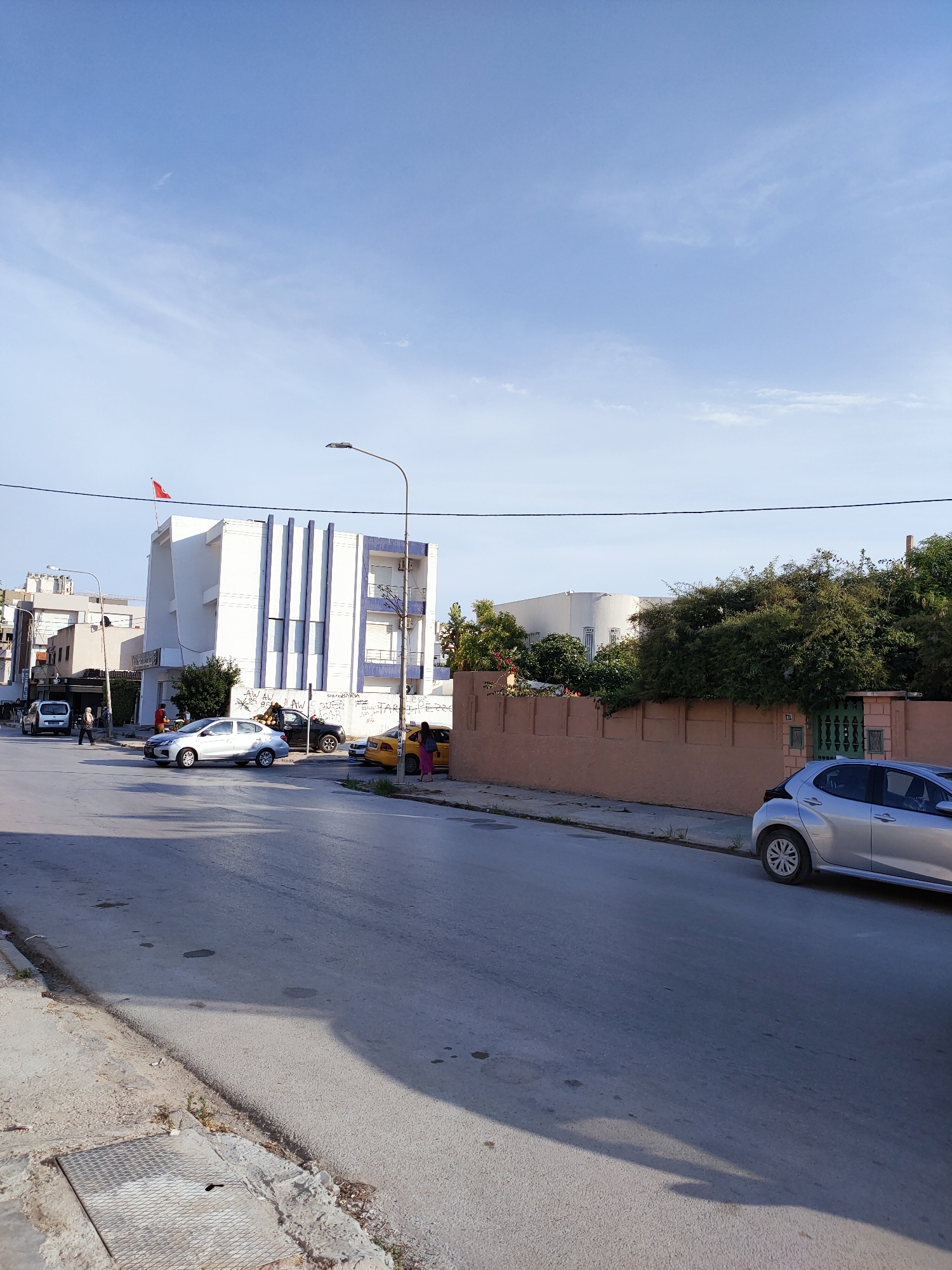 The image depicts a street scene in Ariana, Tunisia, under a clear blue sky with some wispy clouds. The time of day appears to be late afternoon, judging by the shadows cast by the buildings and cars on the road.
In the foreground, a paved street runs diagonally from the bottom left to the top right of the frame. To the left of the street is a sidewalk with a metal grate. A silver car is parked on the right side of the street, its rear end visible. Further down the street, other vehicles are present, including a yellow taxi, a dark car, and a silver sedan. A red flag is visible on top of a white building with blue vertical stripes, suggesting a national or official presence.
Several people are visible in the image. Two individuals are standing near the yellow taxi, and a few more are seen walking on the sidewalk in the middle ground. One person in a hijab is walking near a white van.
The buildings in the background are varied. The prominent white building with blue accents appears to be a commercial or office building. To its left, other structures with a more residential or commercial appearance line the street. On the right side of the image, a long, low, peach-colored wall separates the street from lush green trees and shrubs. Beyond the wall, parts of a white building with a curved facade are visible. Graffiti is present on the peach-colored wall.
The overall atmosphere is one of a typical day in a residential or mixed-use urban area, with everyday activities like driving and walking taking place. The presence of the flag and the distinctive architecture might offer further clues to the specific location within Ariana.