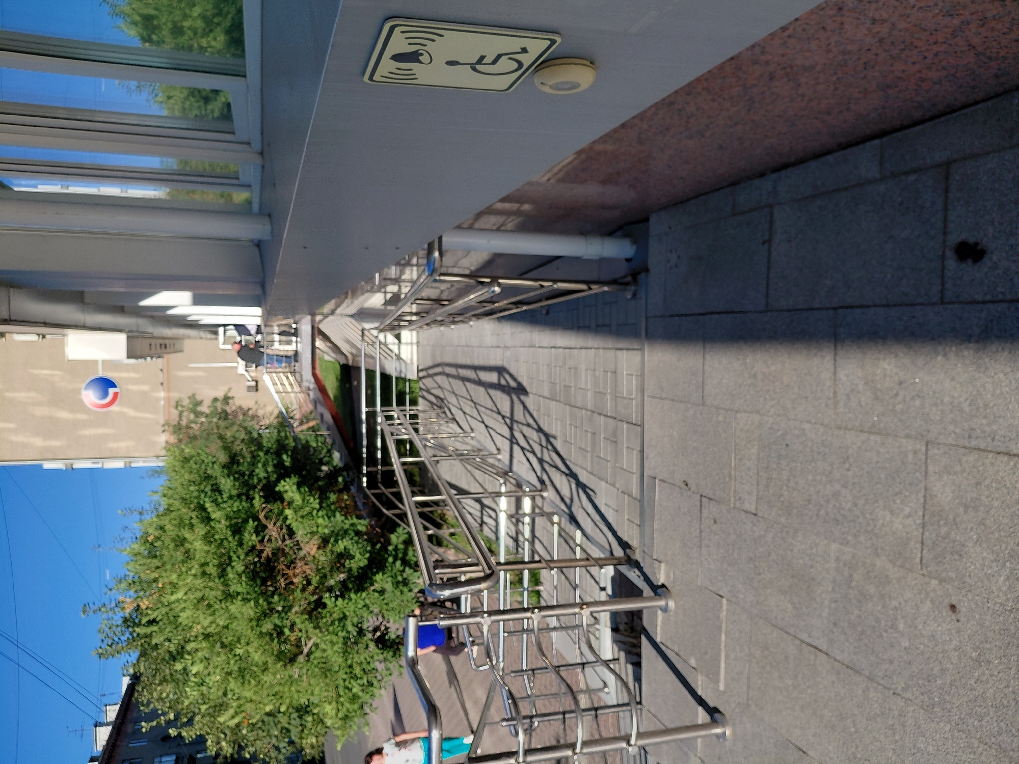 A bright, sunny day is depicted in this medium-angle shot of an outdoor ramp and stairs leading up to a building entrance. The ramp, made of gray pavers, is flanked by a polished metal railing. The building facade is a neutral beige with large, reflective windows. A sign with a wheelchair symbol and a bell, indicating accessibility, is mounted on the wall near a yellow call button. A large green tree partially obscures the view of a residential building in the background, and power lines are visible against a clear blue sky. A woman in a white shirt and blue pants is walking away from the camera on the left side of the frame. Another person in a blue shirt is also visible in the background near the ramp. The scene is located in Novosibirsk, Russia, as indicated by the architectural style and signage.