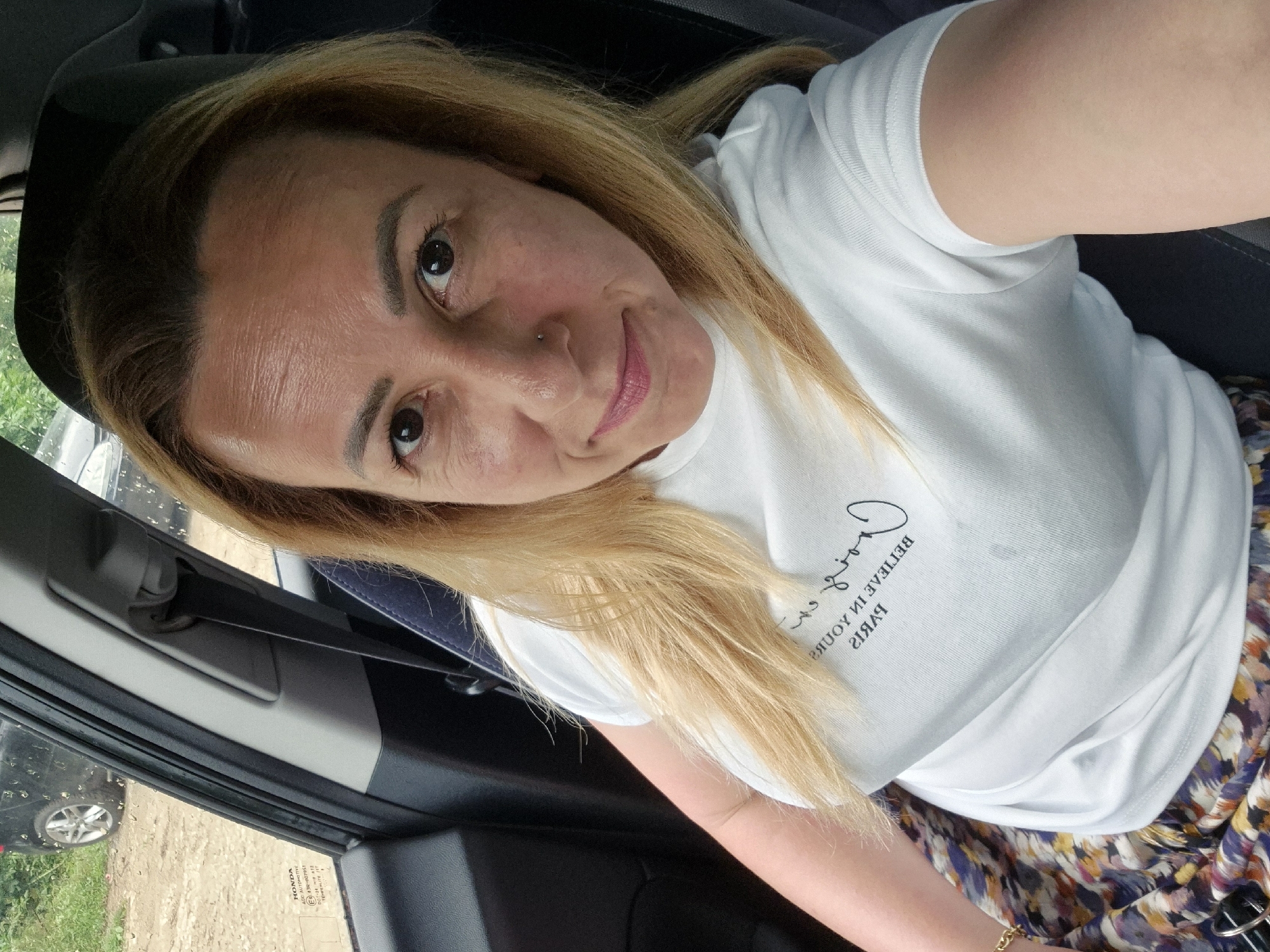 A mid-shot, taken from a slightly high angle, features an adult female seated in the driver's seat of a Honda vehicle. She has long, straight to wavy, light blonde hair and light skin, looking directly at the camera with a faint, closed-mouth smile. Her eyebrows are dark and shaped, and she has a small silver stud piercing on her left nostril (viewer's right) and wears pink lipstick. She is dressed in a white short-sleeved crew-neck t-shirt with inverted black text on the chest that reads "BELIEVE IN YOURSELF PARIS". Below her waist, a patterned garment, possibly a skirt or wide-leg pants, displays abstract designs in shades of blue, purple, yellow, orange, and brown. A gold chain bracelet is visible on her left wrist.
The car's interior shows a dark gray dashboard or door panel to her left, with a gray seatbelt mechanism and strap visible to her right. The A-pillar and window frame are visible to the left. Water droplets are present on the driver's side window. A white sticker with black text is affixed to the lower left of the window, displaying "HONDA", "ACC. AUTOMOTIVE", "E6 43R-000710", "DOT-240 AS3 M18", and "TRANSPARENT TINT". Beyond the window, the exterior background reveals an unpaved dirt or gravel ground surface, with green foliage (grass and trees) in the mid-ground. A dark-colored vehicle, possibly black, is partially visible in the background on the left. The scene is illuminated by natural daylight. Location: Unknown, Unknown.