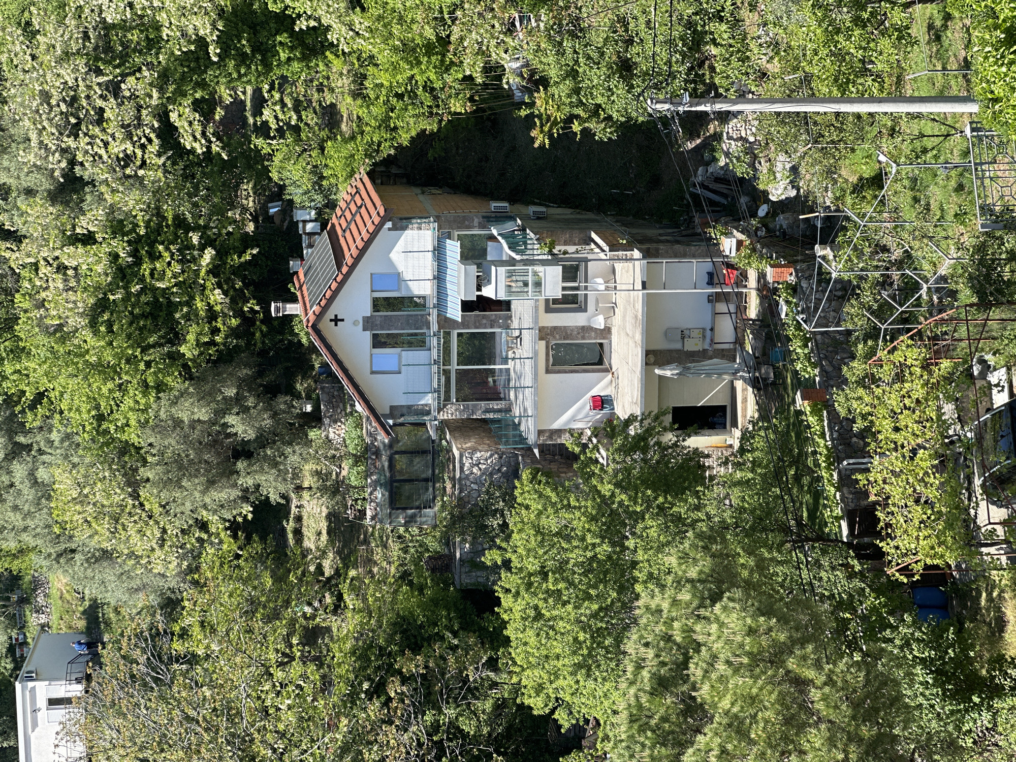 The image depicts a multi-story house nestled among trees, located in or near Budva, Montenegro. The house has a gabled roof with red tiles, multiple windows, and balconies. The facade appears to be a mix of stone and plaster. Electrical wires are visible near the house. The surrounding environment is densely vegetated with trees. A small structure with a wire frame is visible in the foreground to the right of the house. The image suggests the house is located on a hillside or in a wooded area.