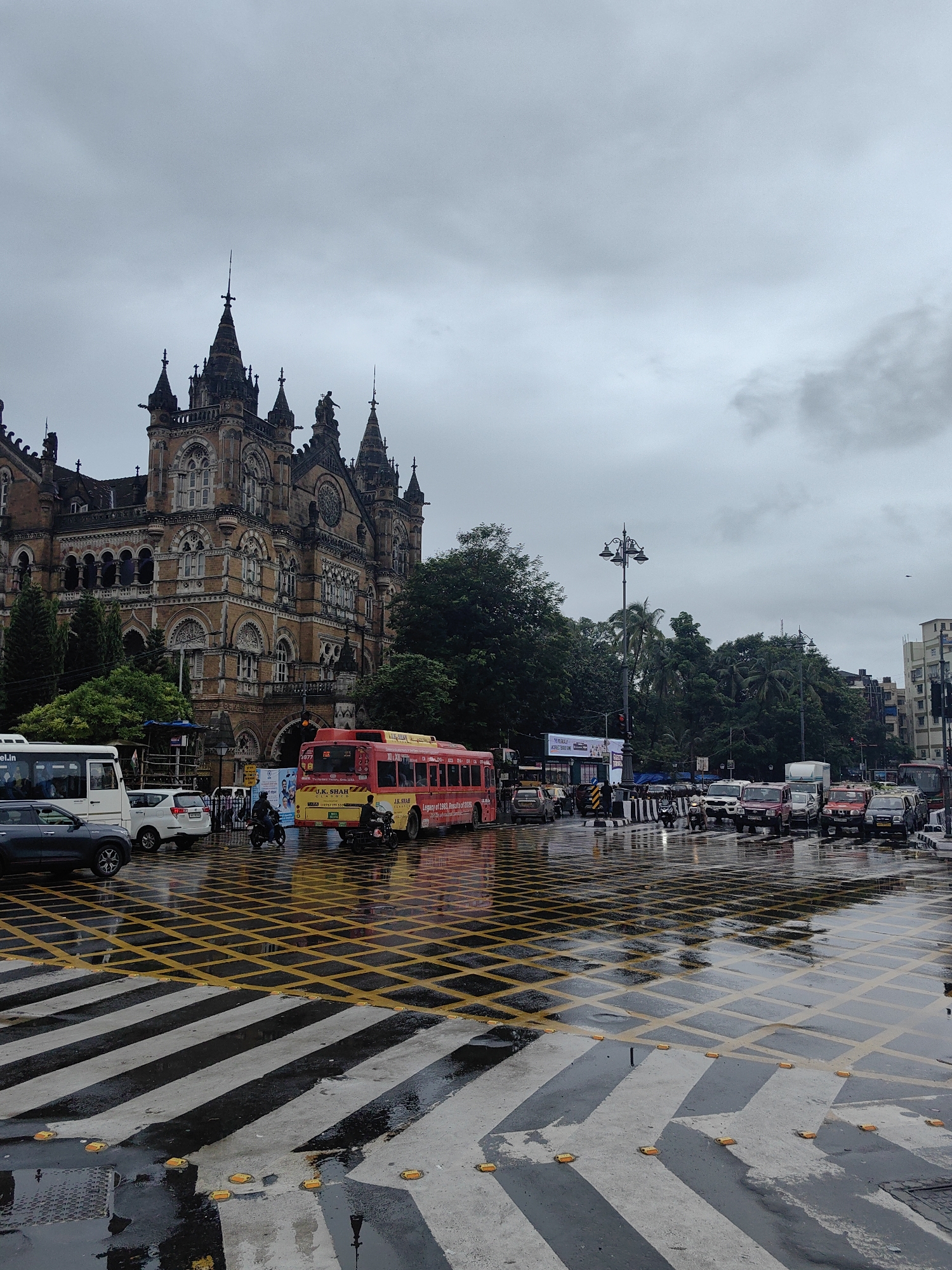 This image depicts a street scene in Mumbai, India, likely in the vicinity of Chhatrapati Shivaji Maharaj Terminus (CSMT), a historic railway station known for its Victorian Gothic architecture. The sky is overcast with heavy, grey clouds, suggesting a rainy or post-rainy day. The street and pedestrian crossing are wet, with reflections visible on the ground, indicating recent rainfall.
In the foreground, a wide pedestrian crossing with black and white stripes and yellow grid markings is prominent. Several yellow road studs are embedded in the concrete. Beyond the crossing, a busy street is filled with vehicles. A red and yellow bus, bearing the branding "J.K. SHAH" and "Legacy of 1982, Results of 2004," is a central focus, stopped at what appears to be a traffic light. Other vehicles, including cars, SUVs, and motorcycles, are also present in traffic.
To the left, a large, ornate, and historic building with multiple spires, arches, and windows dominates the background. This is characteristic of CSMT. Trees and greenery are visible to the right of this building and further down the street, interspersed with modern urban structures like apartment buildings. The overall atmosphere is that of a bustling urban environment during a period of inclement weather.