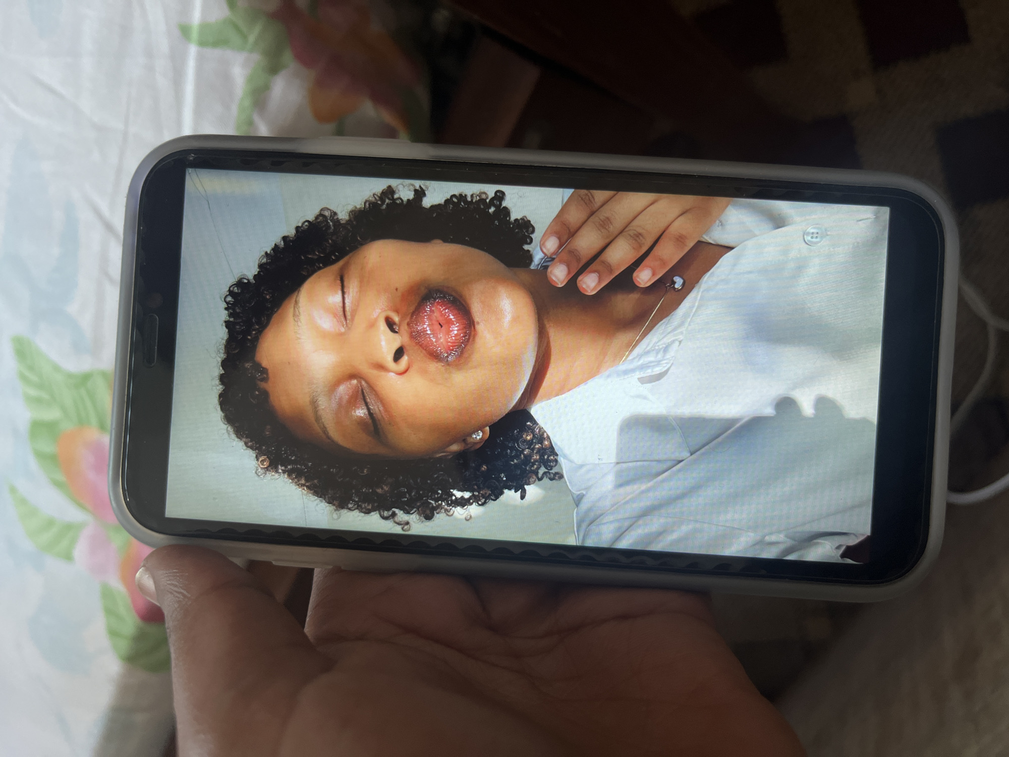 A hand holds a smartphone displaying an image of a person with dark, curly hair lying down, eyes closed, and mouth slightly open. The person wears a light-colored button-down shirt. The smartphone has a light-colored case. The background is out of focus and shows a patterned surface with floral motifs. The location is unknown and is not identifiable as Dar es-Salaam, Tanzania.